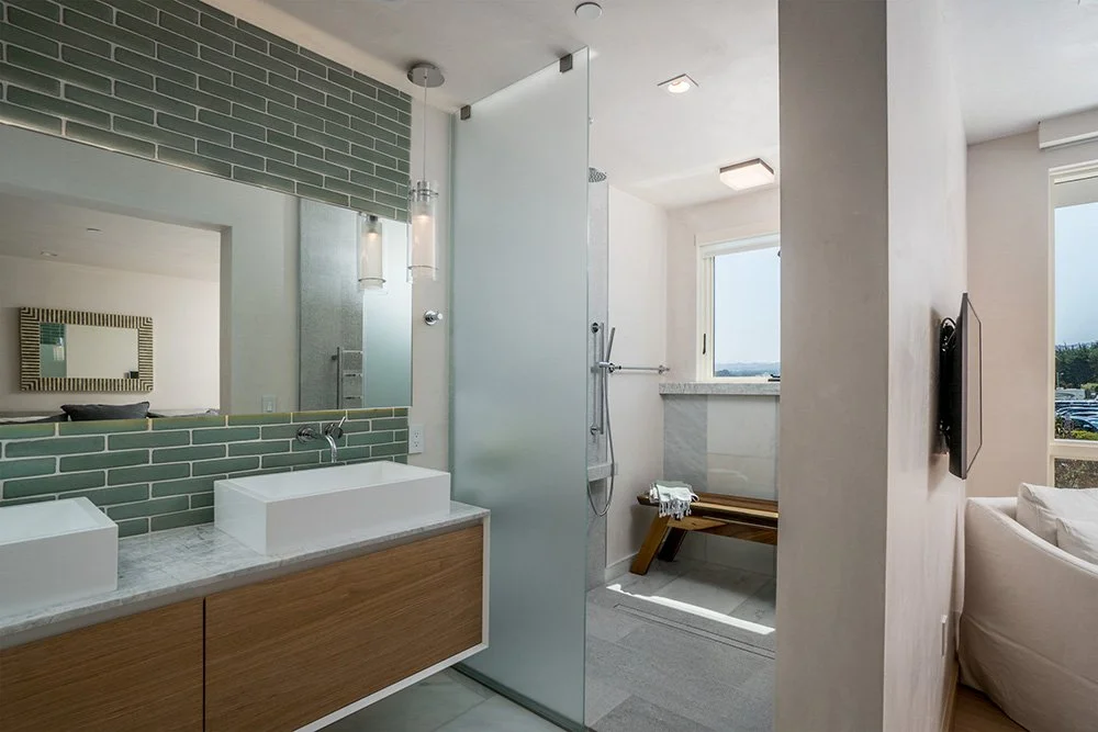 Modern bathroom with green tile backsplash, white vessel sinks, wooden vanity, frosted glass shower door, bench, large window, and a mounted flat-screen TV.