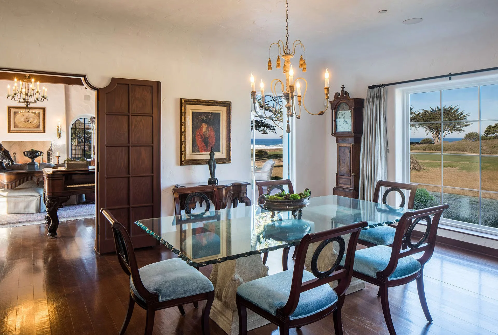 Dining room with wooden table and chairs, large window with an ocean view, vintage chandelier, and artwork on the walls.