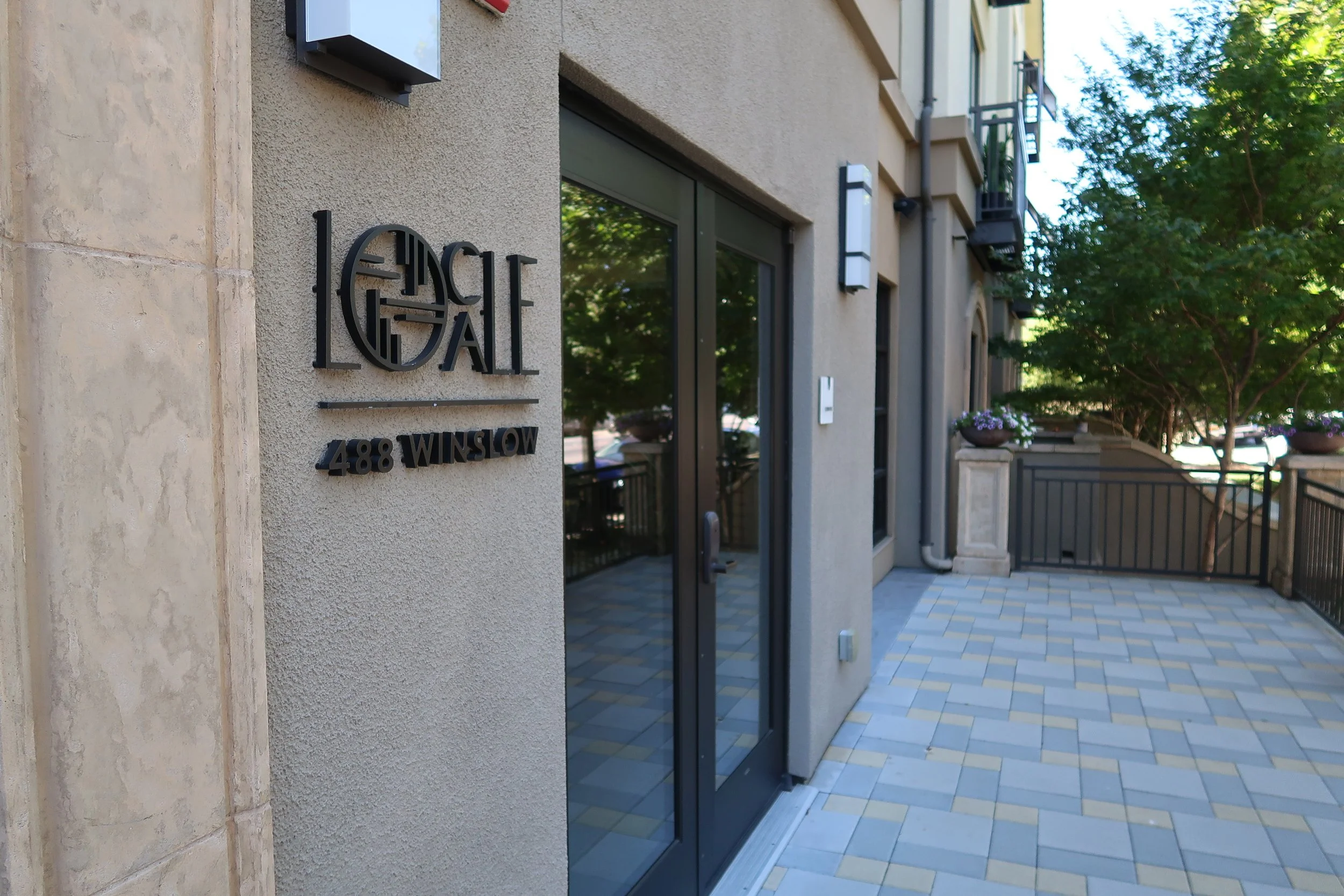 Exterior view of a modern apartment building with a sign reading 'The Local' and address '488 Winslow' on the wall. The building has large glass doors, metal balconies, outdoor lighting, and trees in the background.