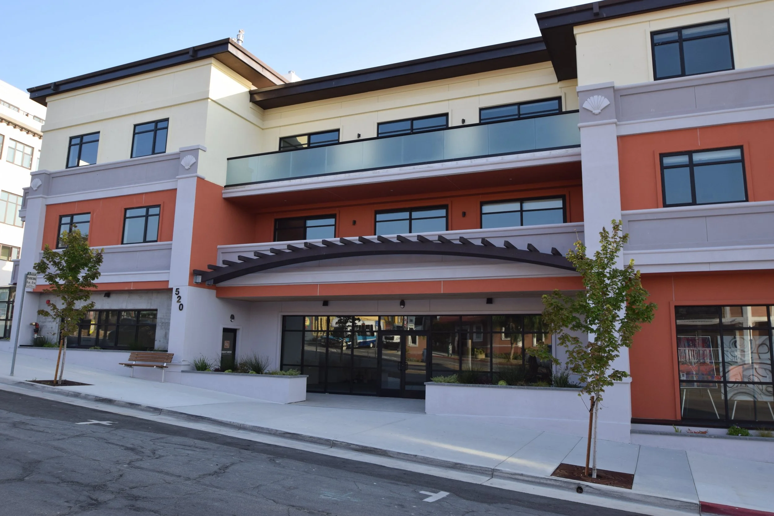 Modern, multi-story building with beige and orange exterior walls, black-framed windows, and a glass balcony. Trees and a bench are in front, with a clear blue sky above.