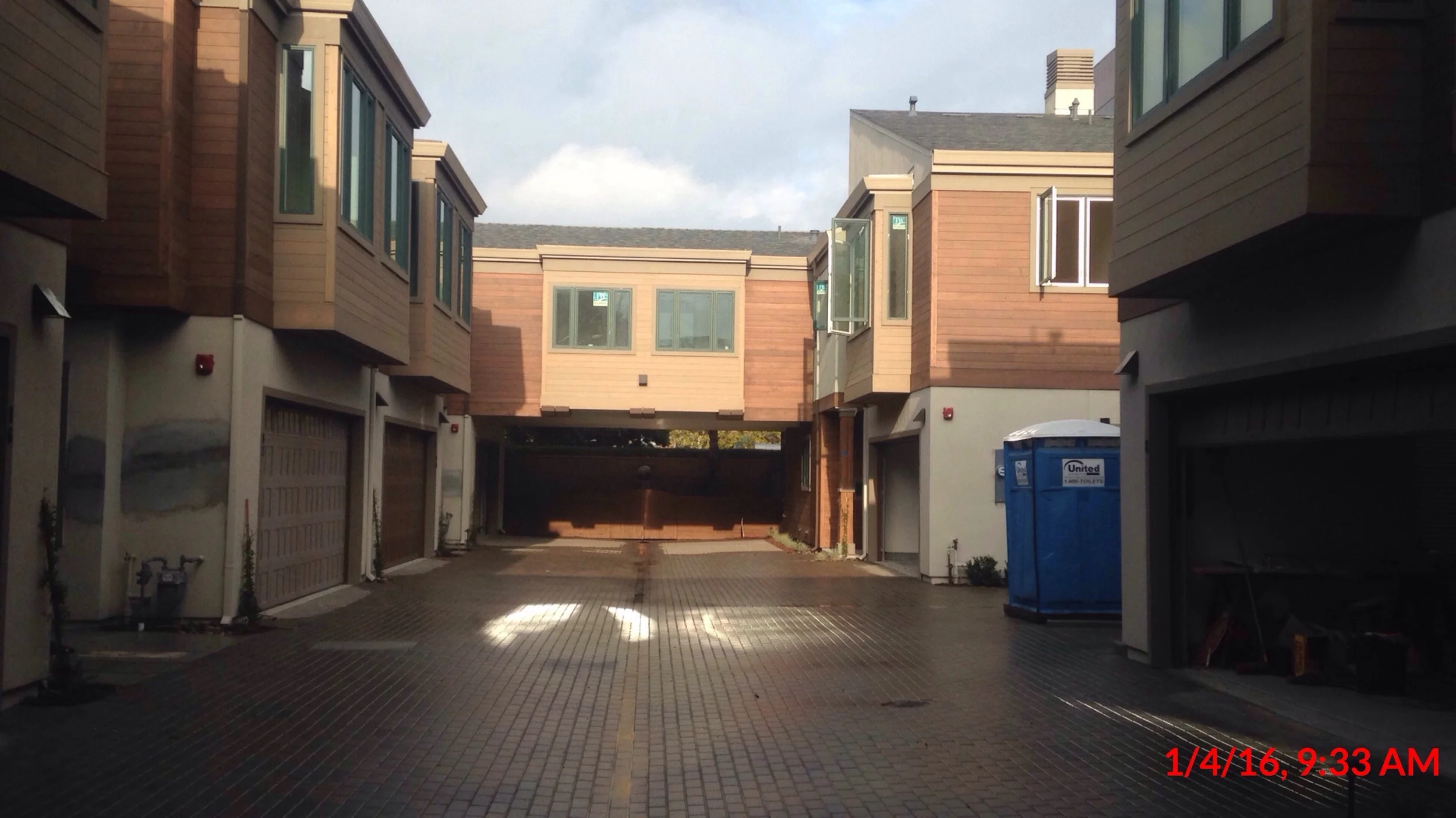 View of a parking area beneath residential buildings with brown and beige exteriors, windows, and a blue trash bin, taken in the morning at 9:33 AM.