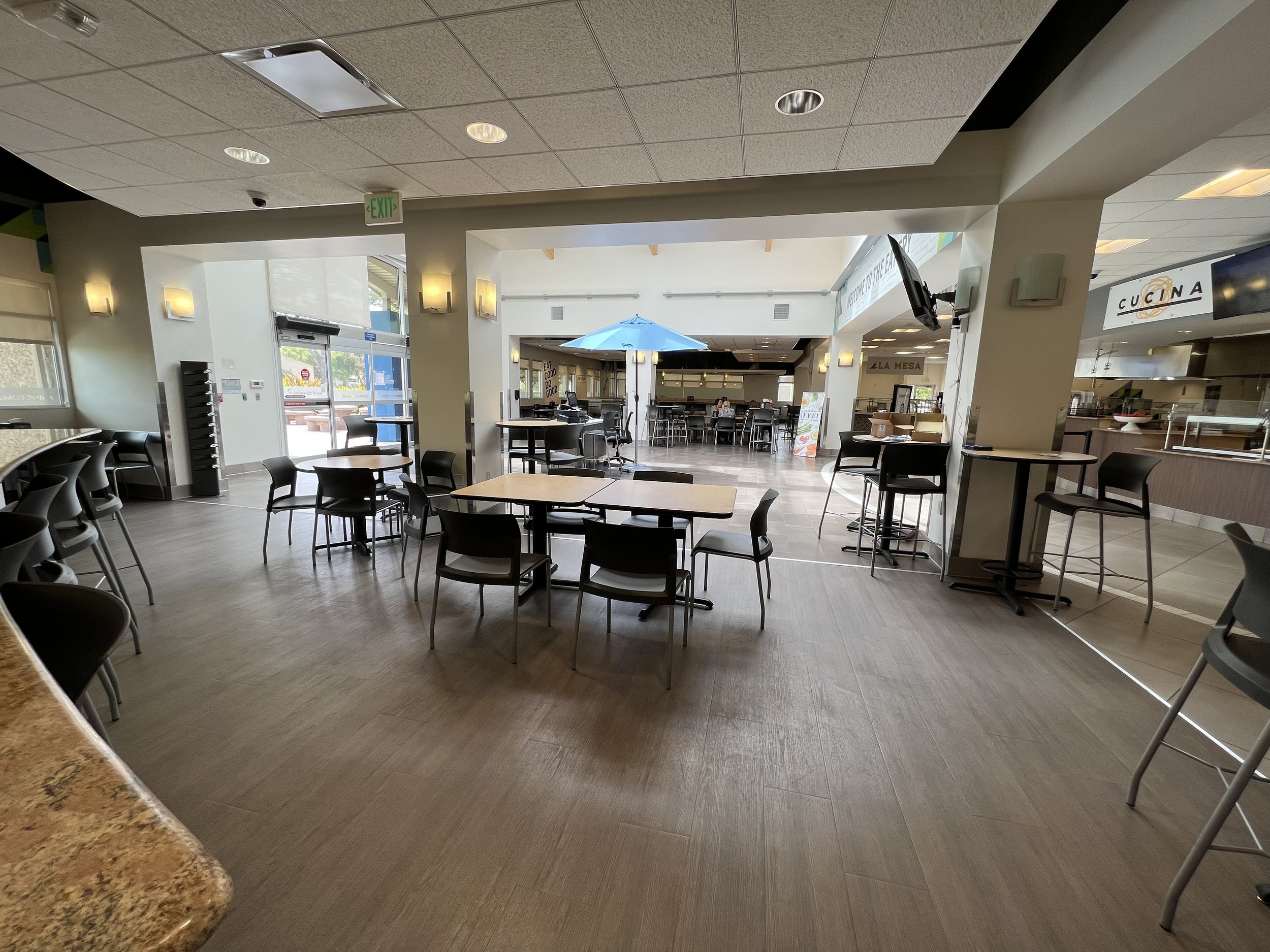 Interior of a casual dining area with tables, chairs, and a counter, with a view of the outside through large glass windows and an open kitchen area in the background.