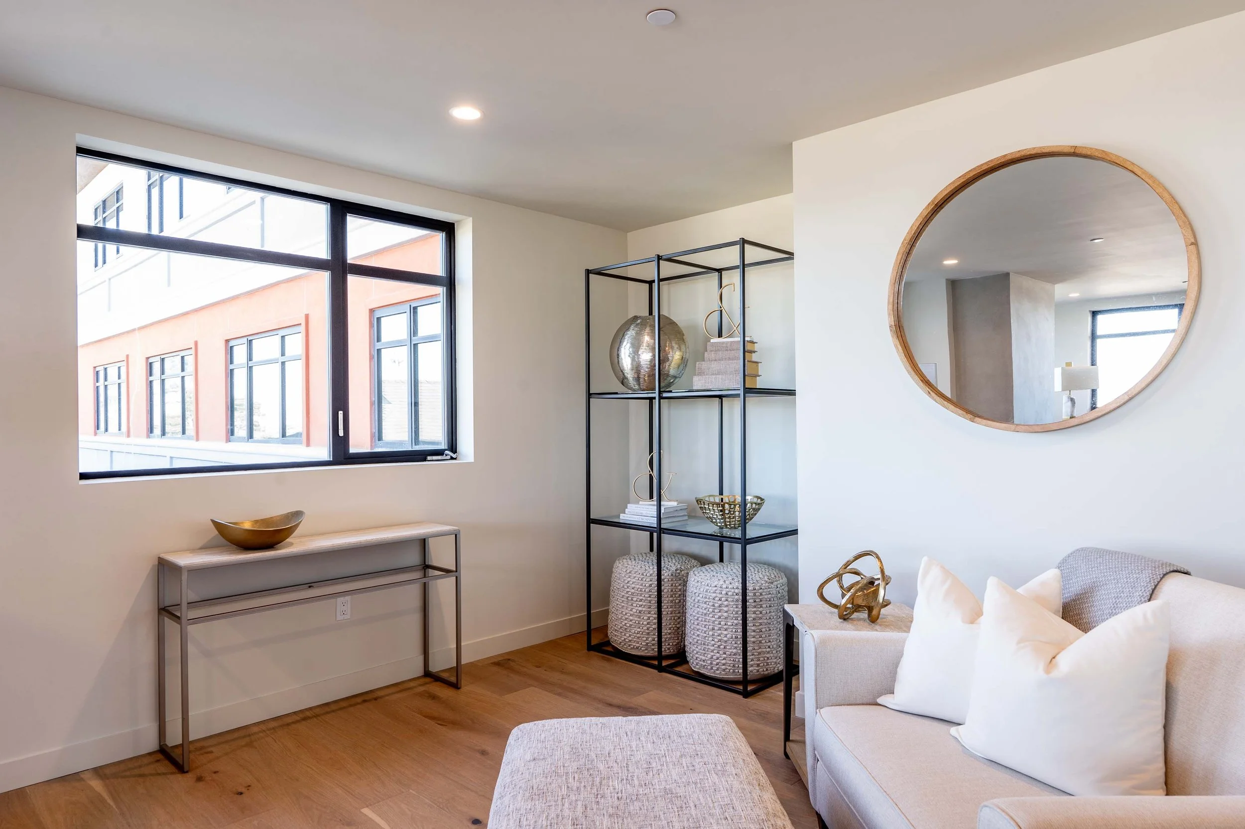 A modern living room with a large window, a round wall mirror, a black metal shelving unit decorated with a large metallic vase, books, and a decorative bowl, a white armchair with pillows, a beige ottoman, and a console table with a wooden bowl on i