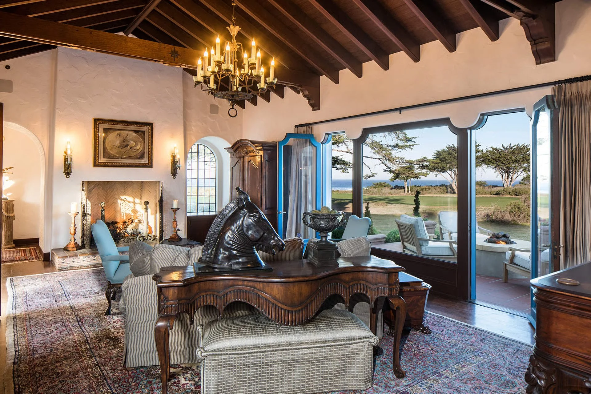 Living room with a fireplace, chandelier, and large windows showing trees and ocean view.