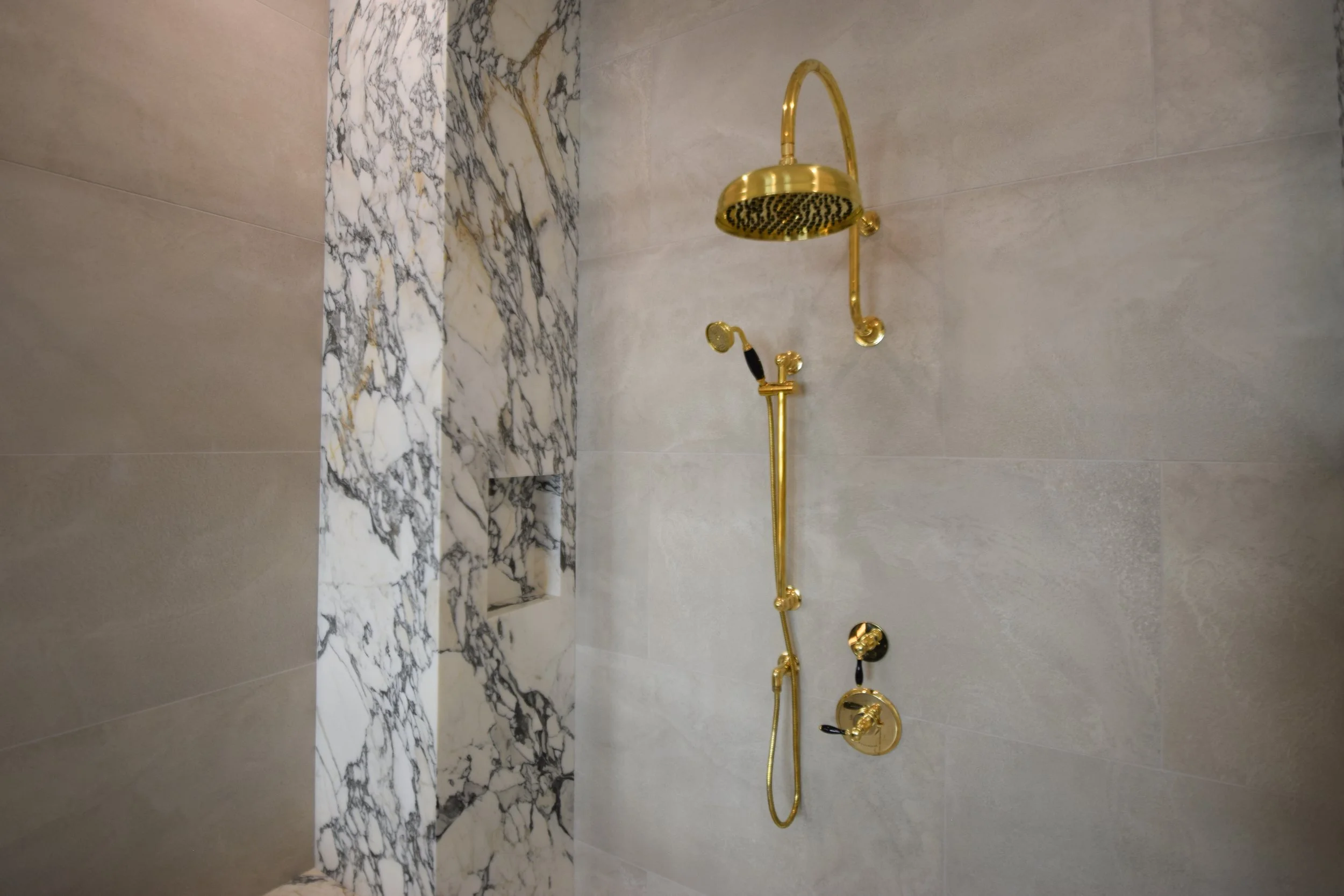A bathroom shower area with a gold-colored showerhead, control knobs, and handheld shower. The wall has beige tiles and a section of marble with white, gray, and black veining.