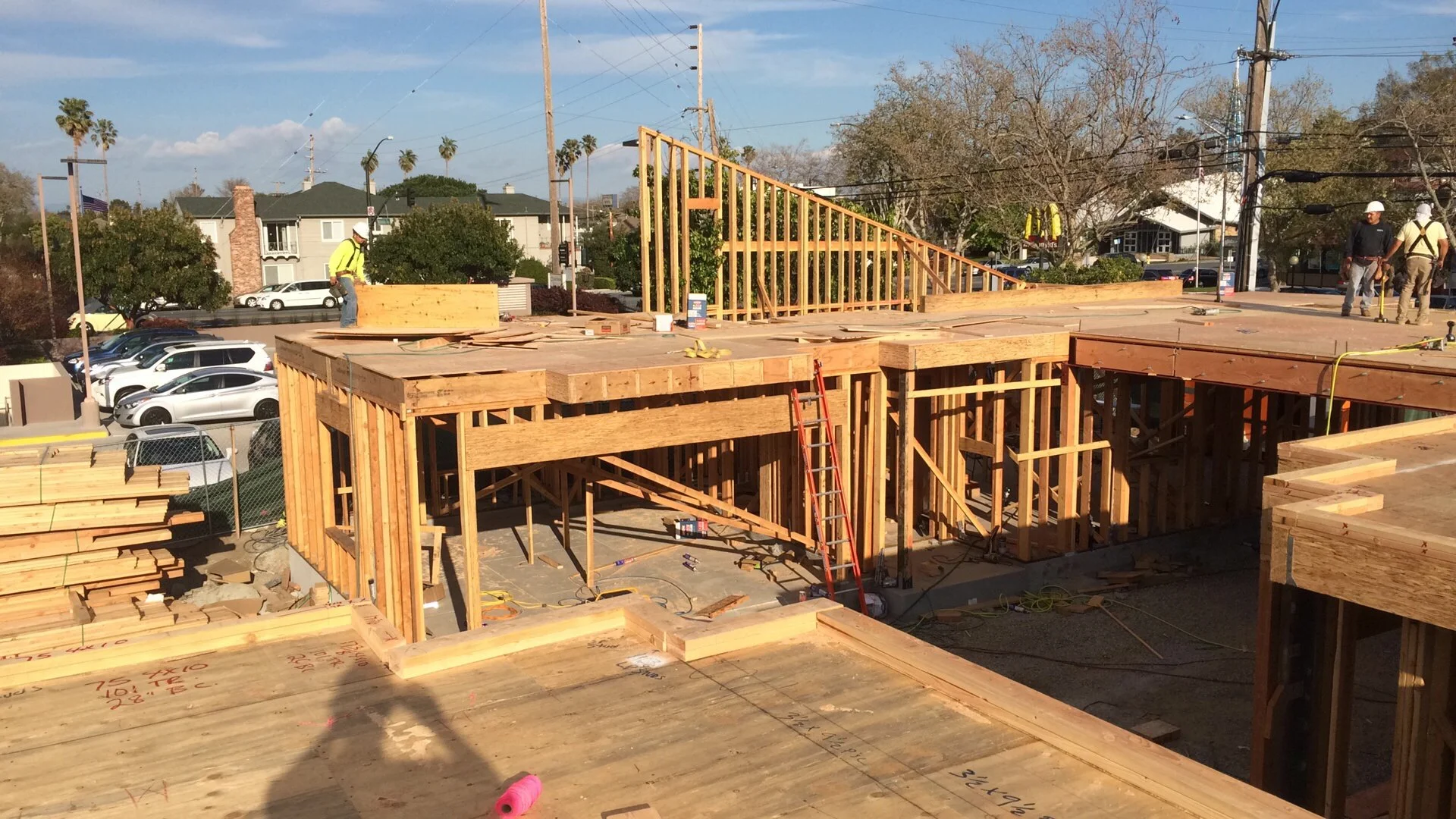 Construction site with wooden framing for a building, workers with safety helmets, and parked cars in the background.