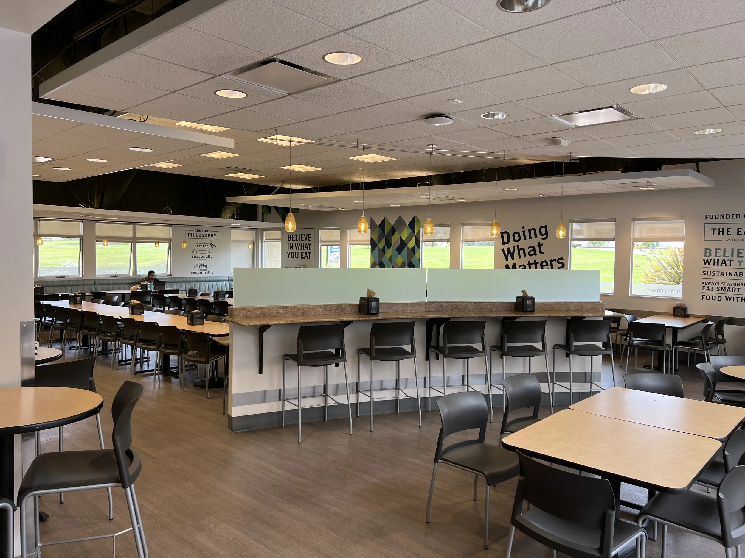 Empty dining area in a modern, well-lit cafeteria or restaurant with round tables, black chairs, large windows, and decorative wall signs.