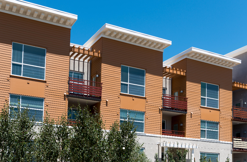 Modern apartment building with a wooden exterior, large windows, and small balconies, set against a bright blue sky.