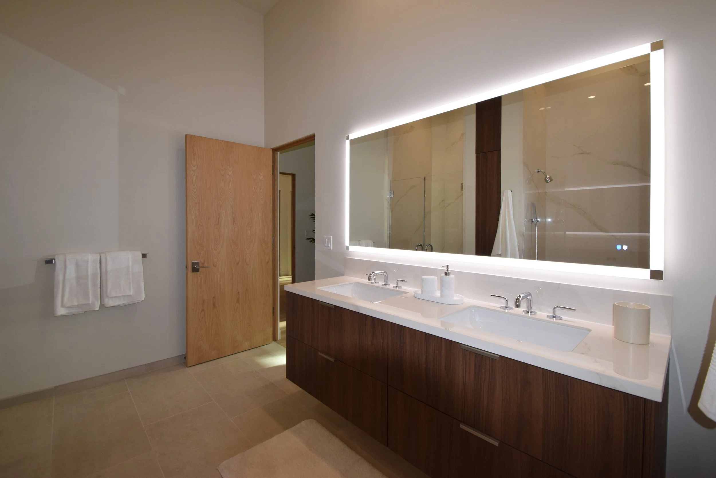 Modern bathroom featuring a large backlit mirror above a double sink vanity with a wooden base, beige tiled floor, and a wooden door. Towel rack with white towels on the left wall, and a shower area with a glass panel and white towel hanging inside.