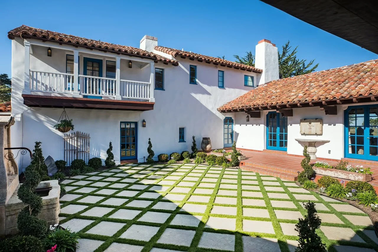 Sunny courtyard with white stucco house, red tile roof, blue window frames, potted plants, lush green lawn with grid pattern, and blue sky.