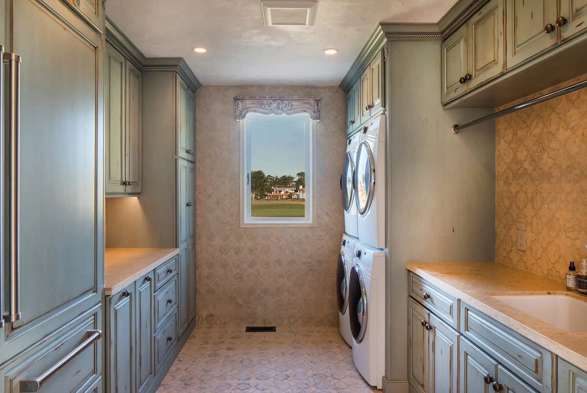 A laundry room with light green cabinetry on both sides, a window at the end showing an outdoor view, and a washer and dryer set under the window. The room has beige countertops and patterned beige flooring.