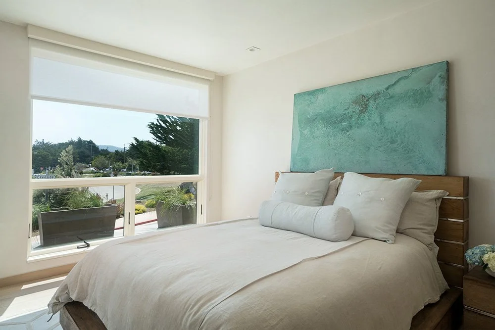 A bright bedroom with a large window, a white bed with pillows, a blue abstract painting above the bed, and a nightstand with flowers.