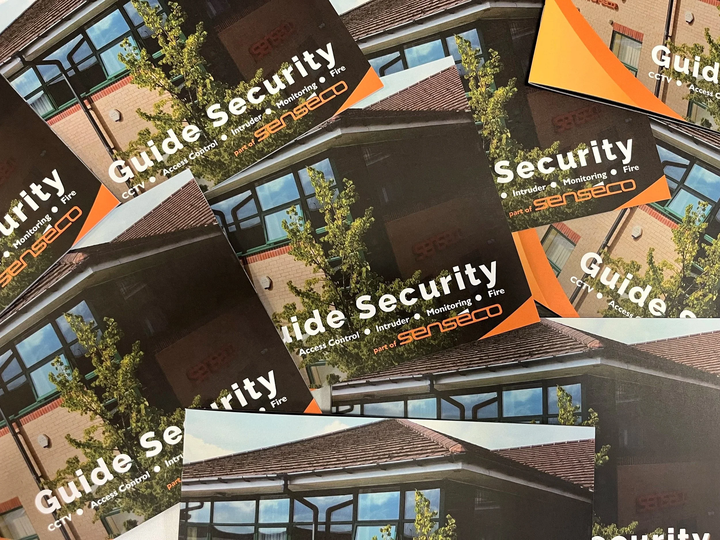 Multiple copies of a security guide brochure. The cover shows the exterior of a building with large windows and a red-tiled roof, some green trees, and a partly cloudy sky. The title reads 'Guide to Security' with subtitles 'Access Control, Intruder,