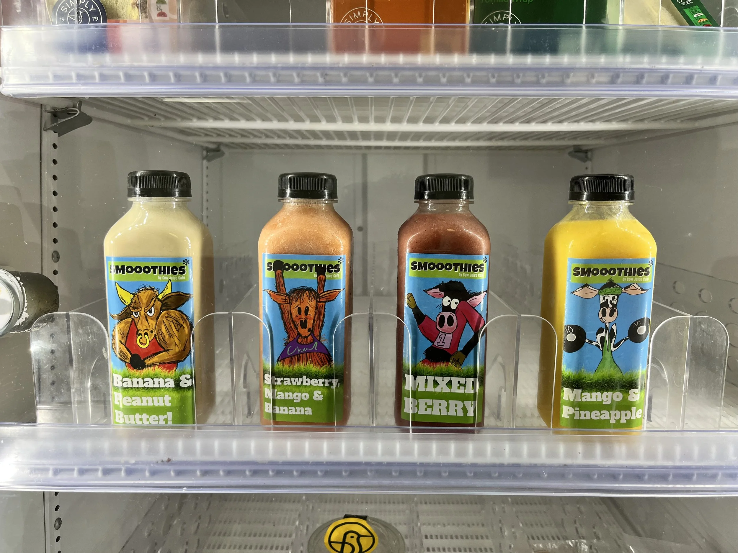 Four bottles of smoothie drinks in a refrigerator, labeled with cartoon animal characters and flavor descriptions: Banana & Peanut Butter, Strawberry, Mango & Banana, Mixed Berry, Mango & Pineapple.