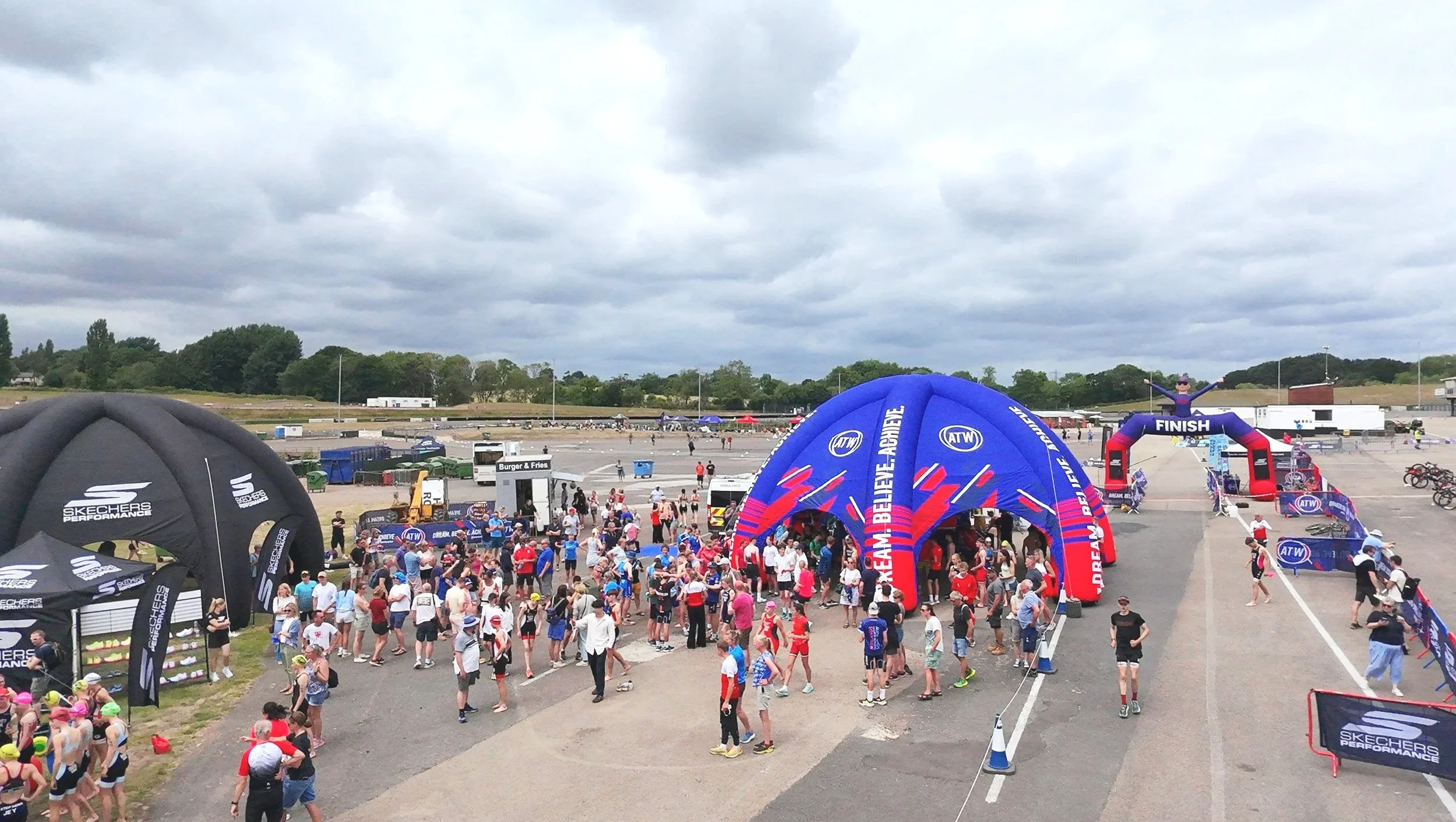Crowd gathered at an outdoor athletic event starting line with inflatable arches labeled 'FINISH' and 'ATW'. People, some wearing caps and sportswear, gather around tents and staging areas on a cloudy day.