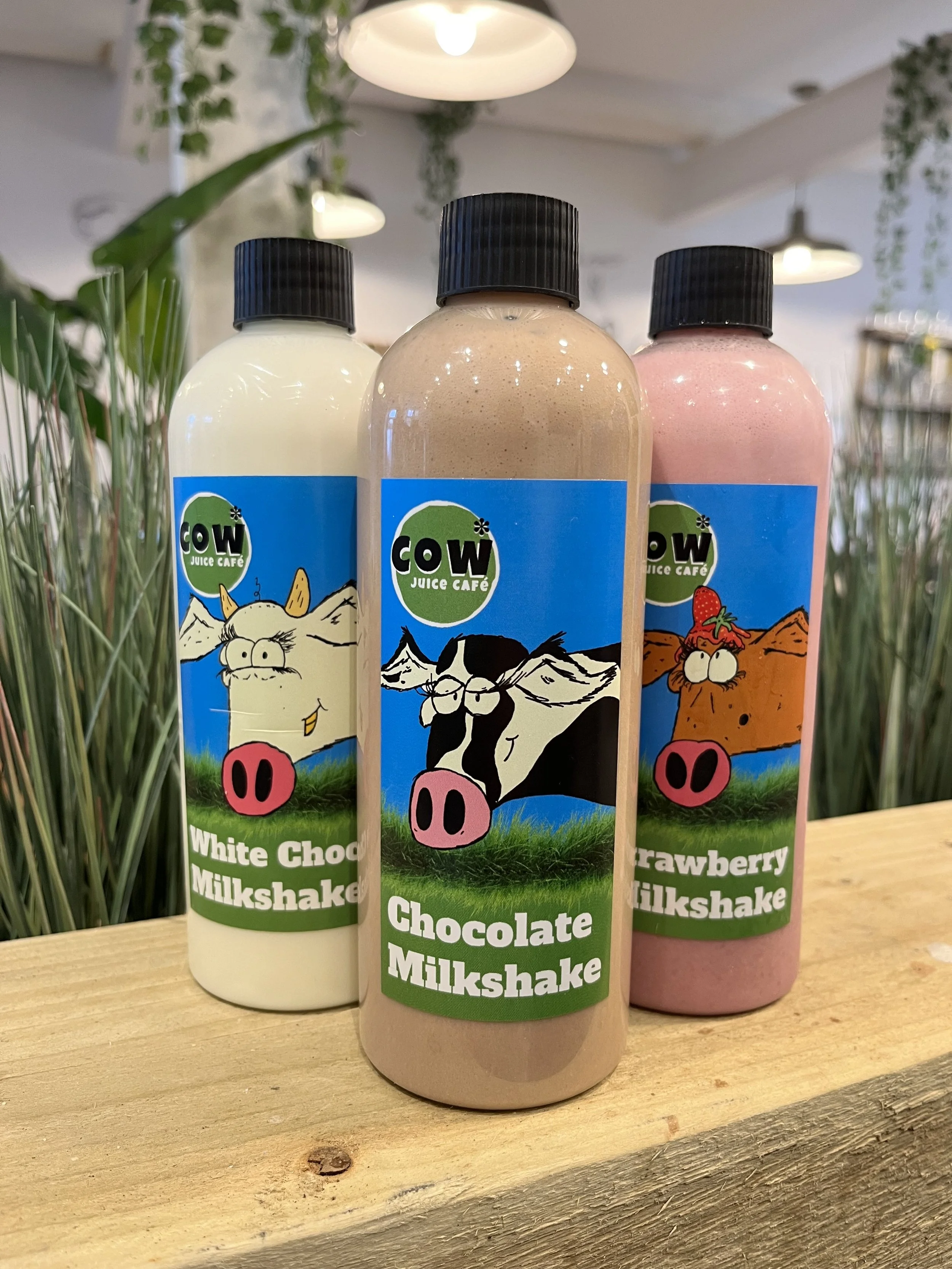 Three bottles of milkshake on a wooden surface with cafe decor background, featuring cartoon animals with glasses and pink pig noses on the labels for different flavors: white chocolate, chocolate, and strawberry.