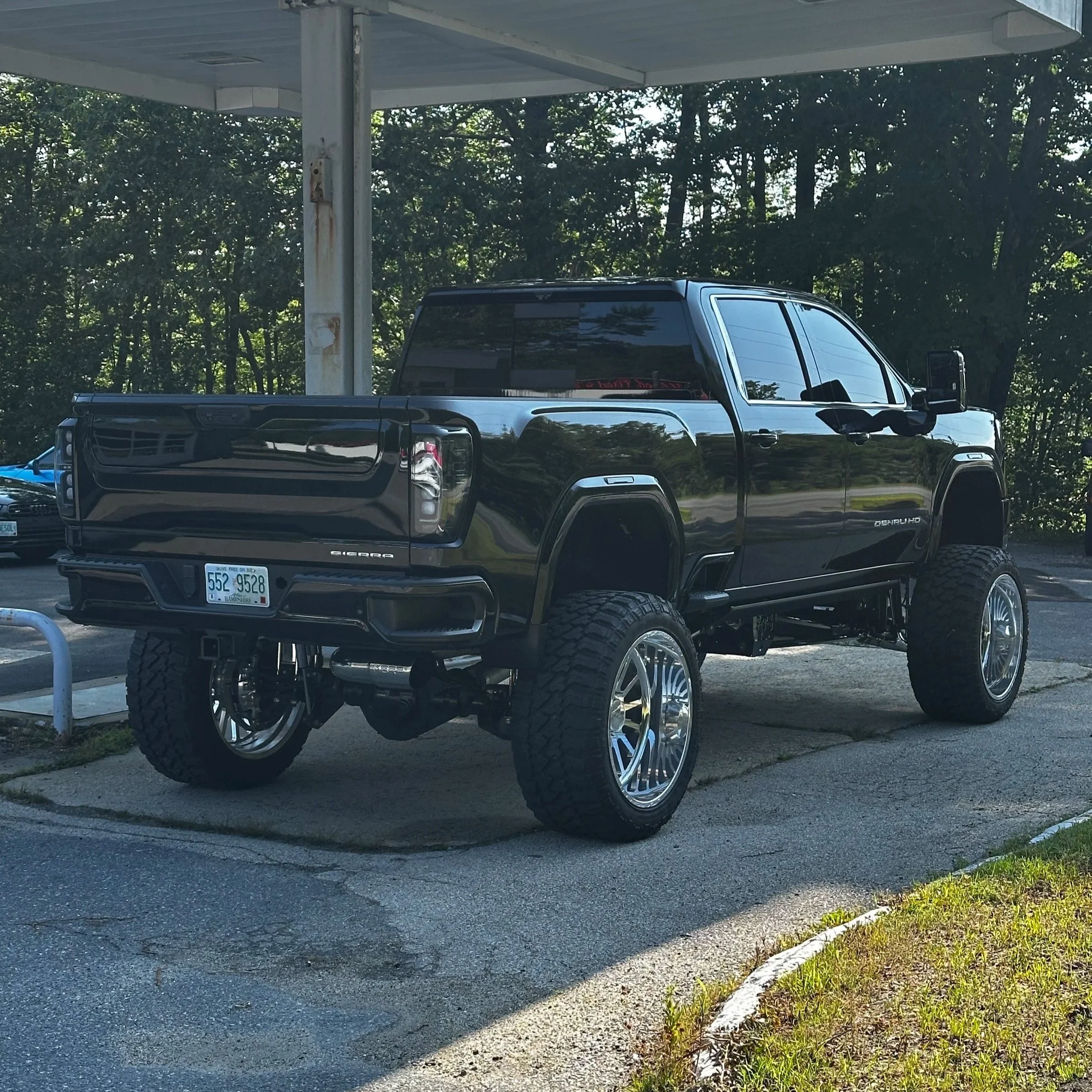 Black lifted pickup truck with large wheels parked under a carport with trees in the background.