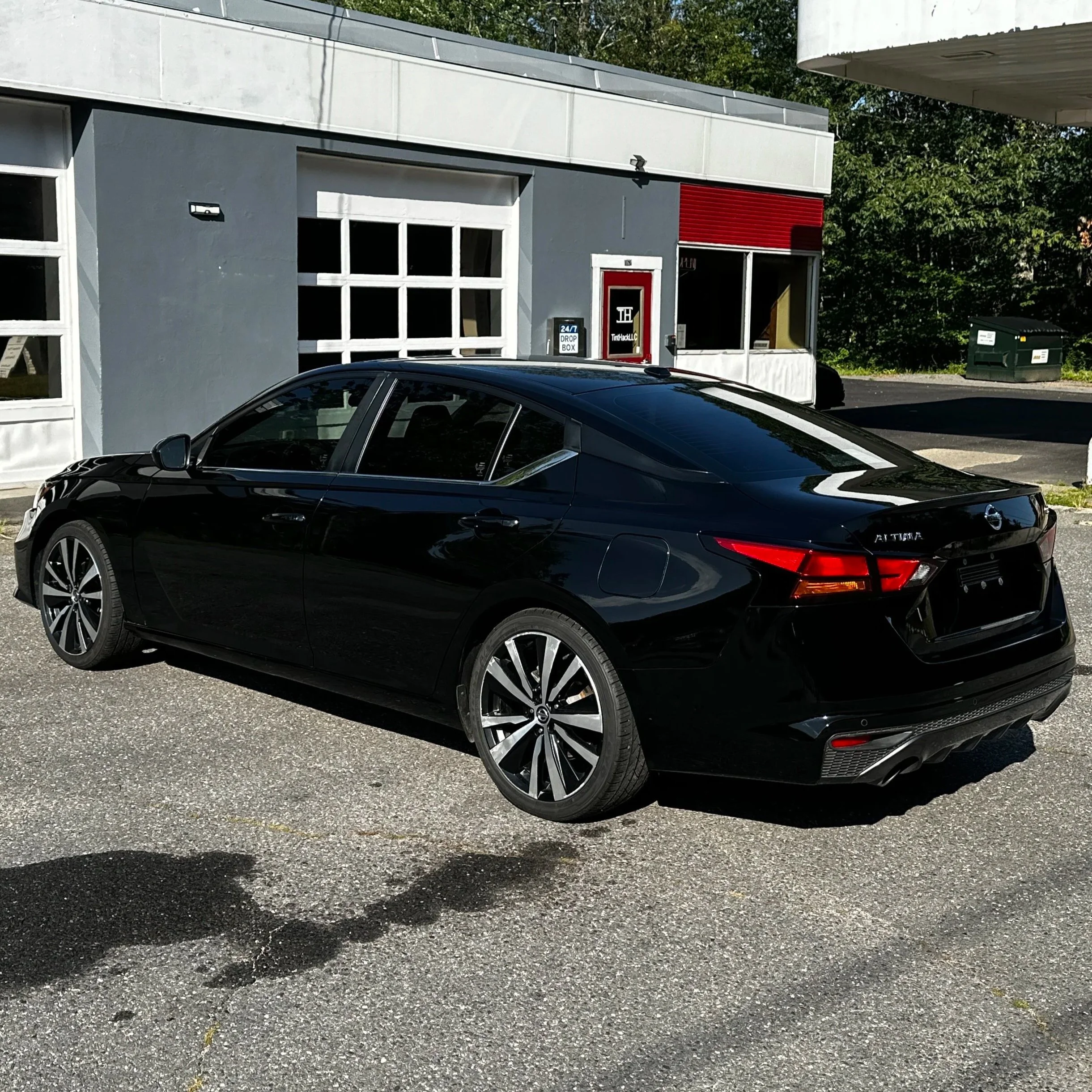 Black Nissan Altima sedan parked outside a building with white and gray exterior walls, large garage-style window, and a red accent wall.