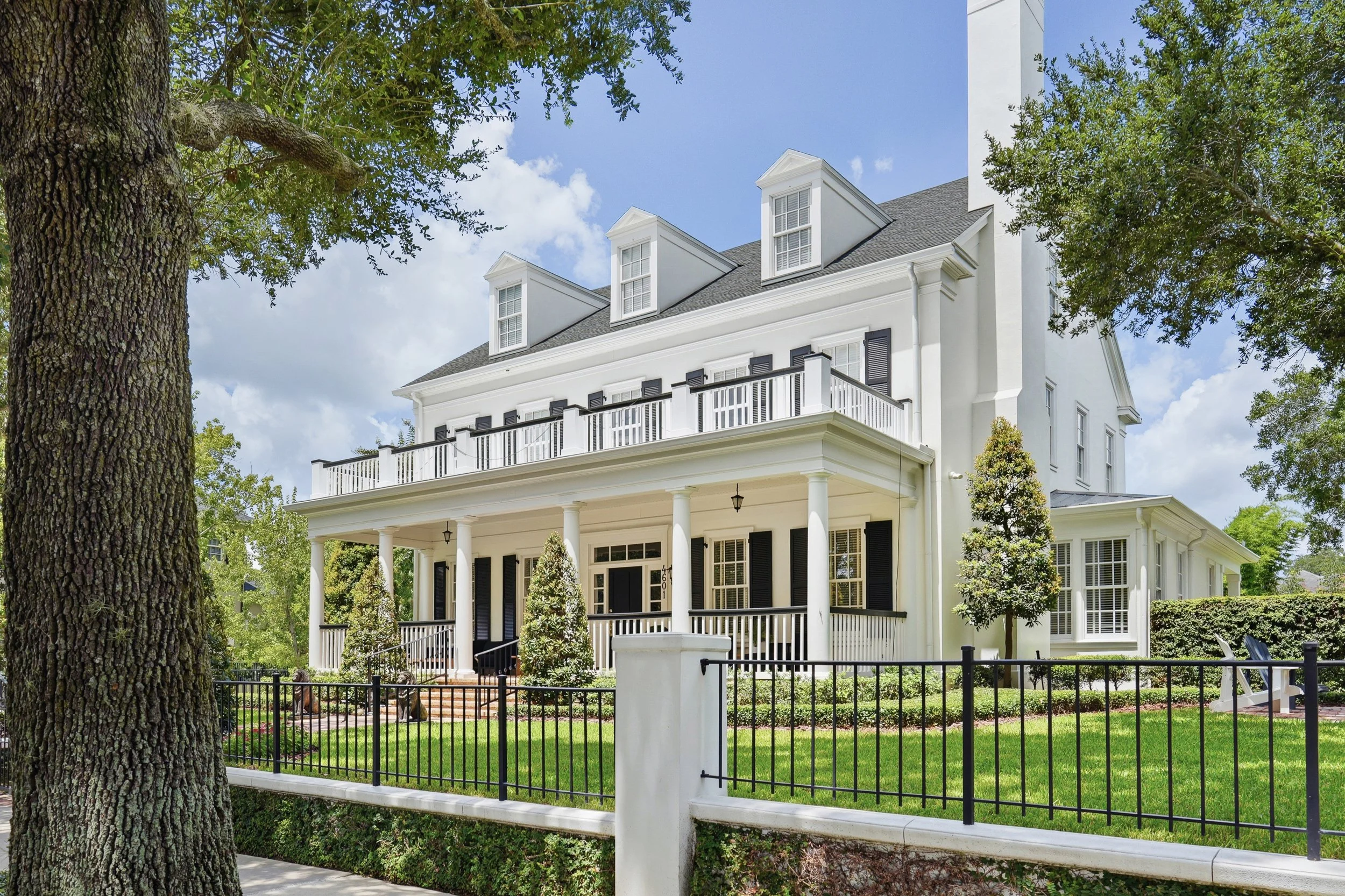  Classic architectural elegance framed by mature oaks, featuring a gracious front porch, second-floor balcony, crisp white façade and timeless black shutters. 