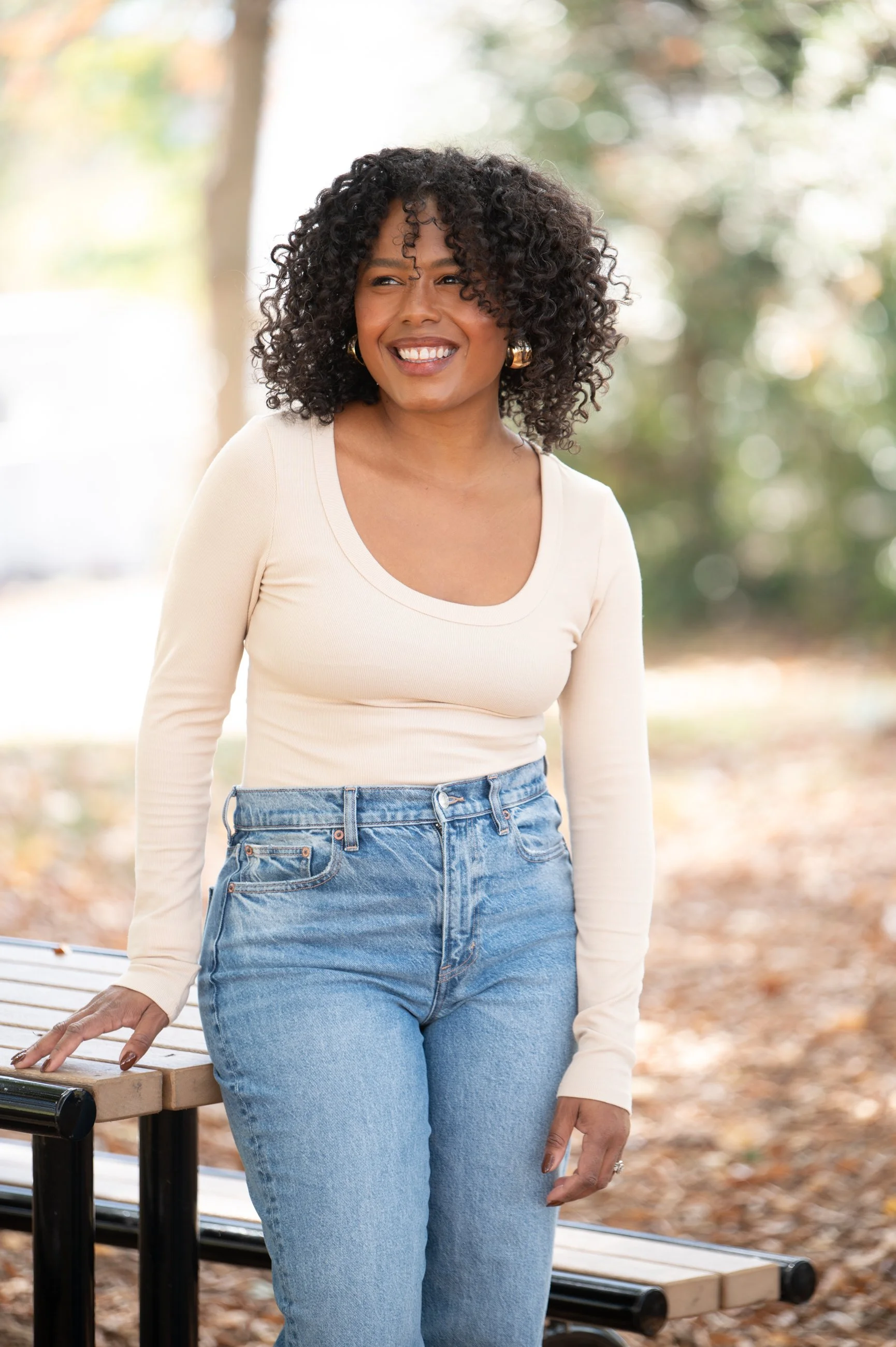 Caroline in jeans and beige long sleep scoop neck shirt leaning against picnic table with right hand gently placed on top of picnic table
