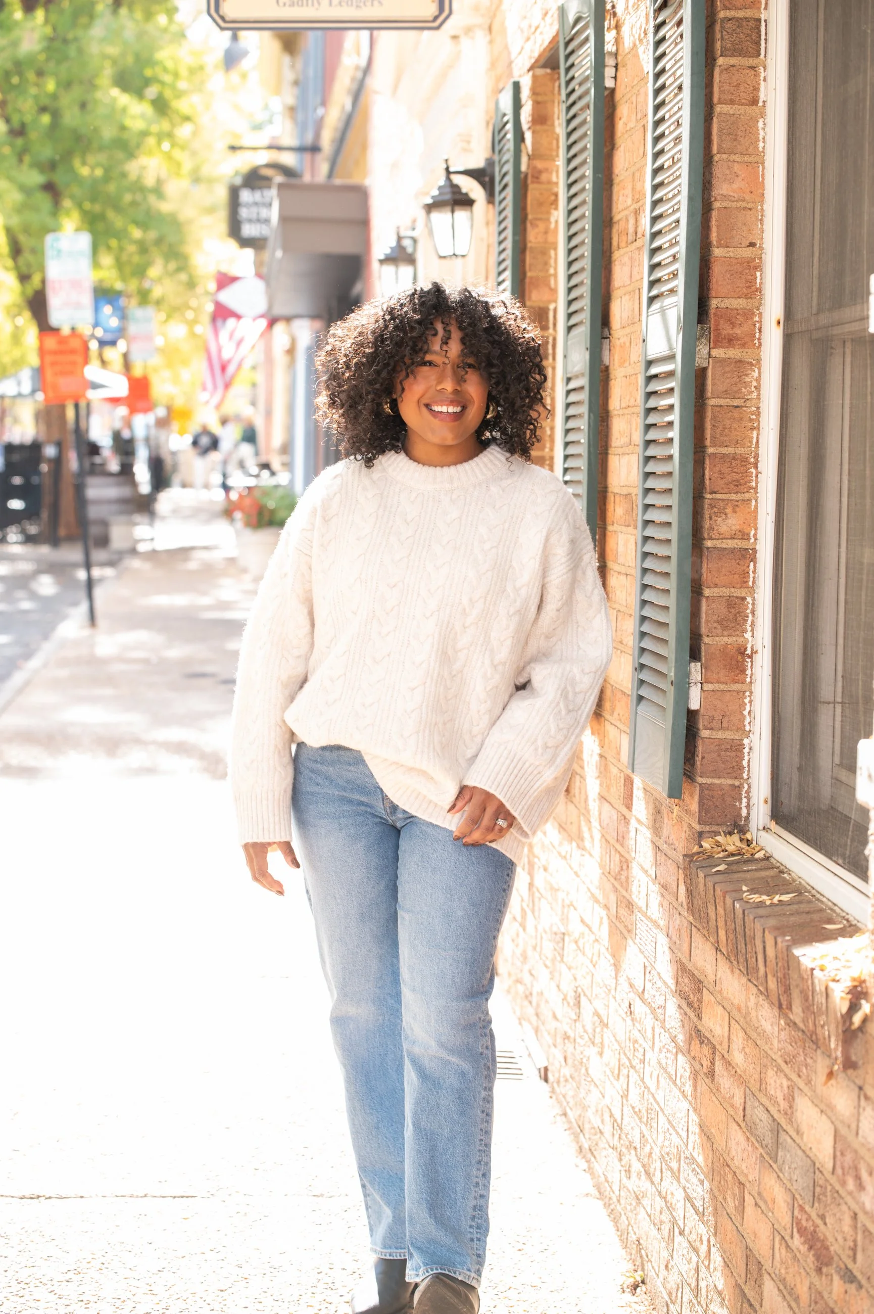 Caroline wearing jeans and white sweater leaning against brick building
