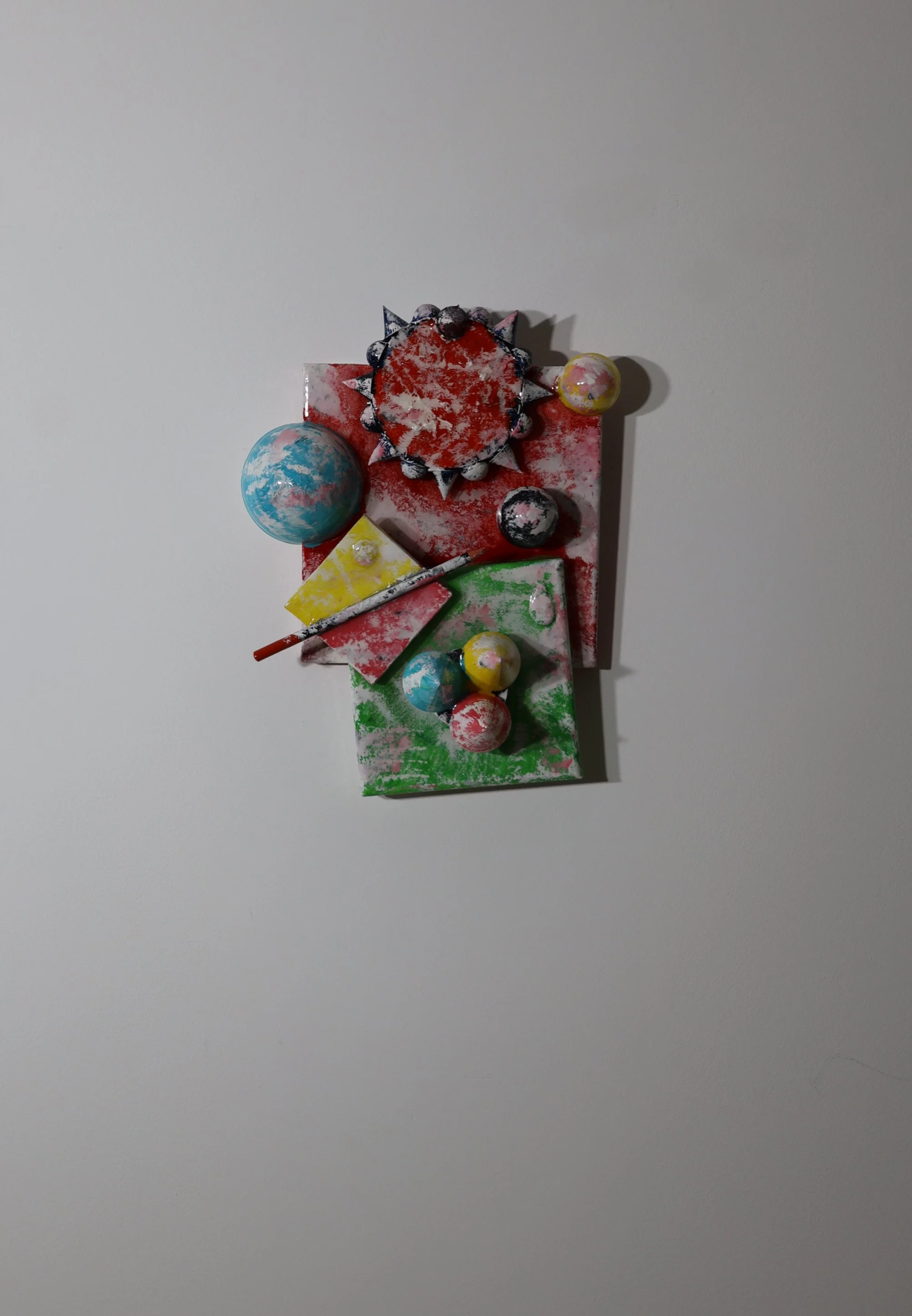 Colorful abstract art installation on a white wall featuring painted spheres, geometric shapes, and structures in red, green, yellow, black, and multicolor with splattered paint effects.