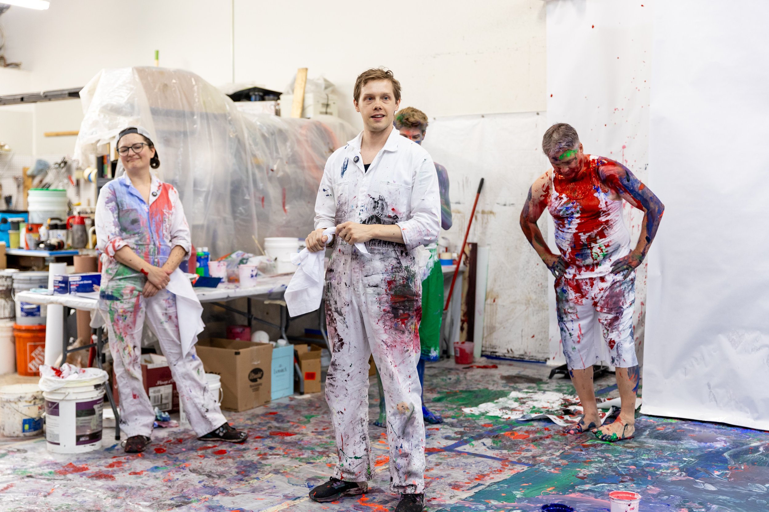 Three people stand in a room with paint splattered coveralls and a messy dropcloth on the floor