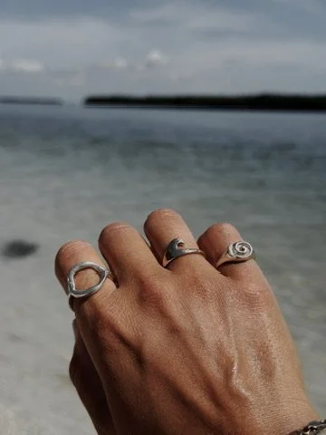 A hand with multiple rings and bracelets is raised against a beach background with blurred ocean and surf