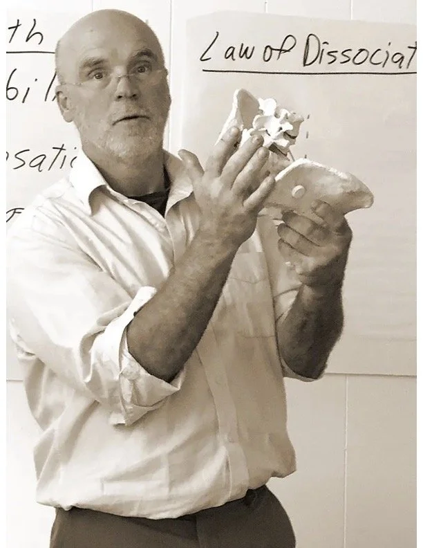 A man with glasses and a white shirt holding a skull model, standing in front of a whiteboard with handwritten notes about the "Law of Dissociation." Massage CE Instructor