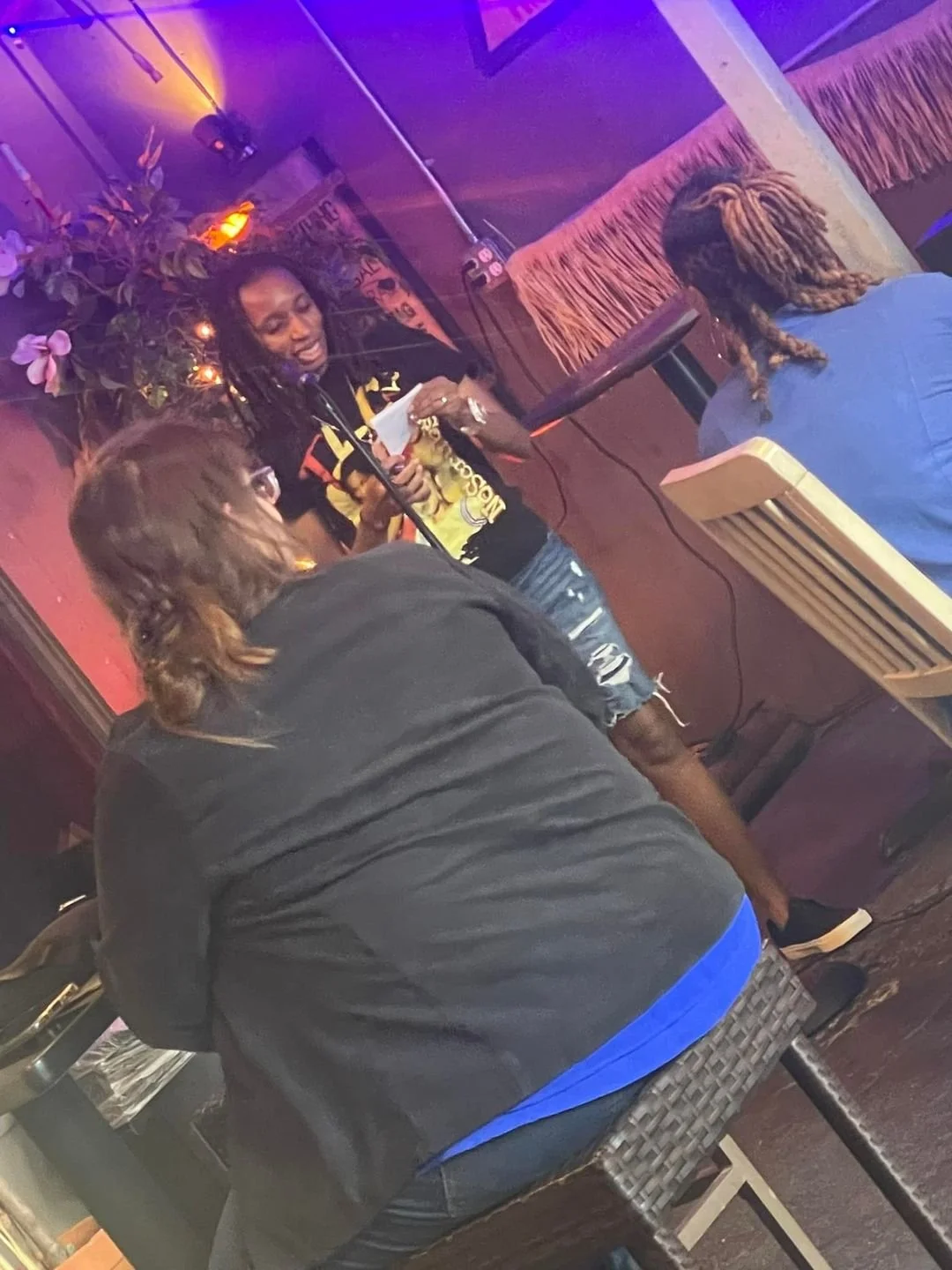 A woman with dreadlocks and a colorful shirt reads from paper at a microphone in a dimly lit venue with purple and orange lighting; two women with long hair sit nearby, one with back facing camera and the other with side profile.