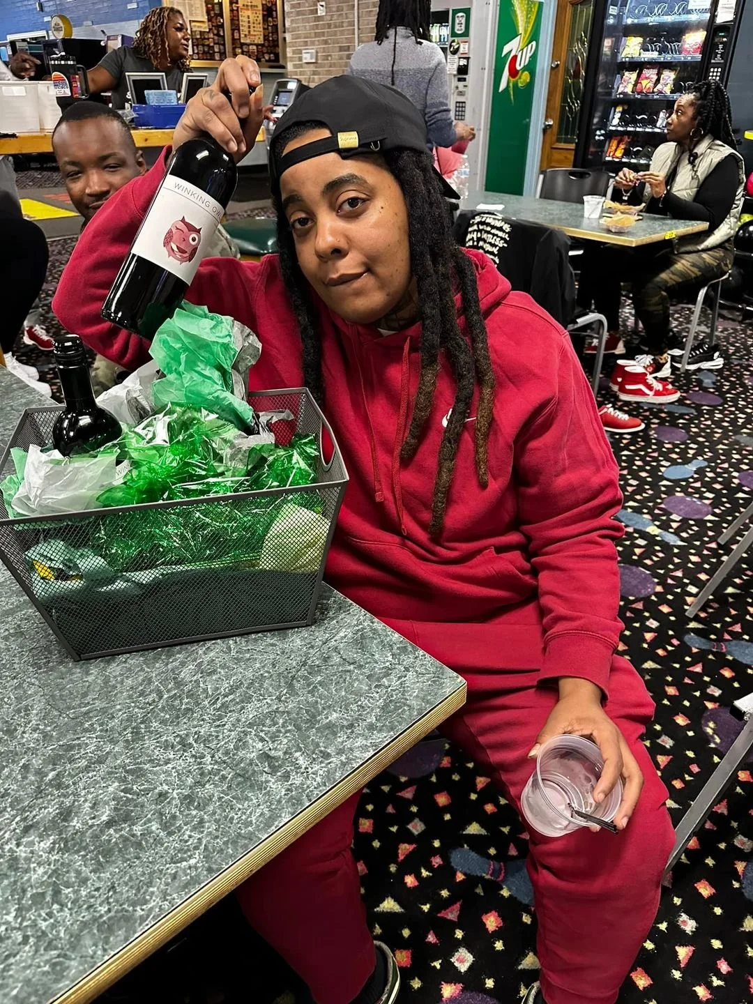 Person wearing a red hoodie and matching pants, sitting at a table in a casual restaurant or gaming area, holding a plastic cup with a straw. They are also holding a bottle labeled with a pig cartoon and the words 