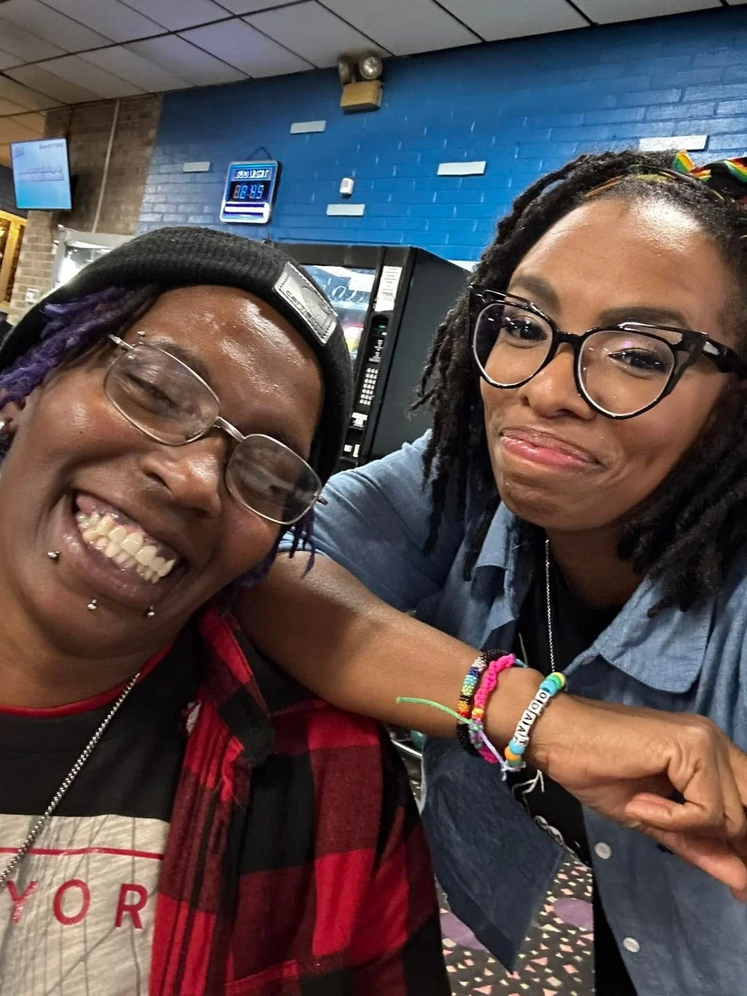 Two smiling women with glasses taking a selfie inside a fast food restaurant, with a blue brick wall and a vending machine in the background.