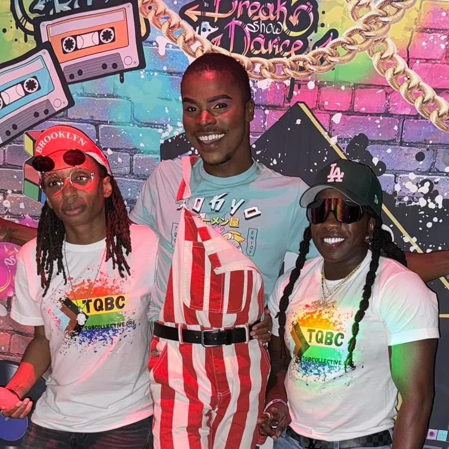 Three people standing in front of colorful graffiti with a mural that says 'Dance' and 'Break'. Two women on either side wear white T-shirts with a colorful 'TQBC' logo. The woman on the left wears glasses, a red Brooklyn cap, and has dreadlocks. The