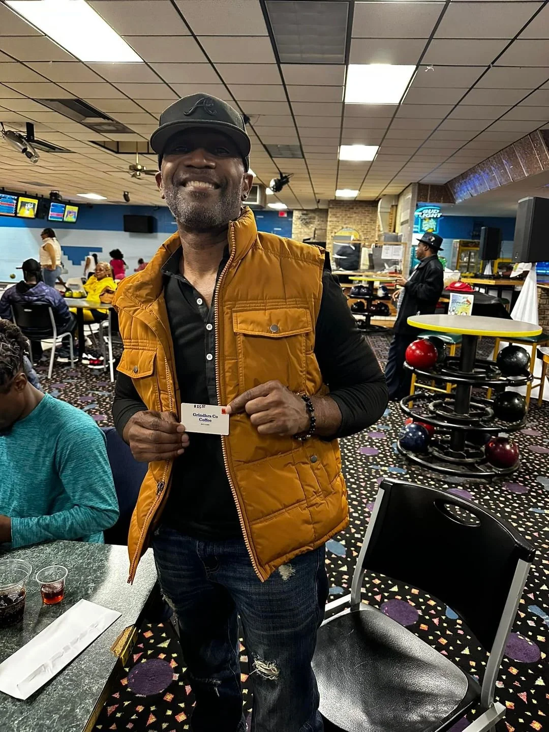 A man smiling and holding a card at a bowling alley, with people in the background and a bowling ball rack nearby.