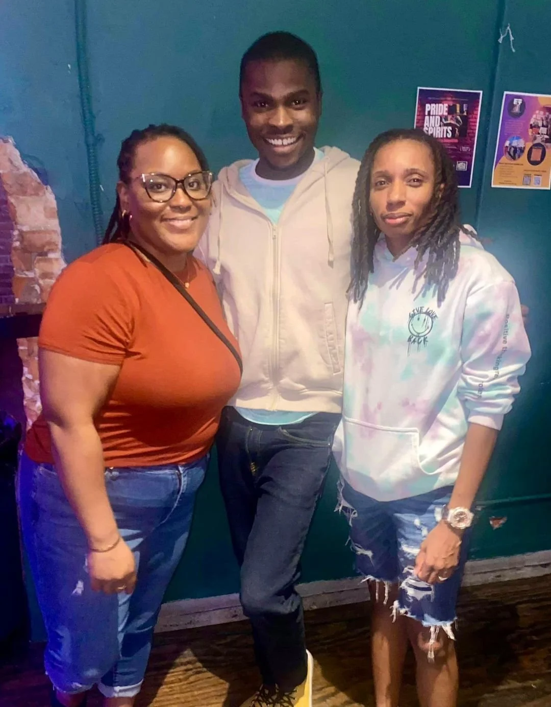 Three people standing together indoors, smiling at the camera. The woman on the left is wearing glasses, an orange shirt, and blue jeans. The man in the middle is wearing a white hoodie and dark jeans. The woman on the right is wearing a tie-dye hood
