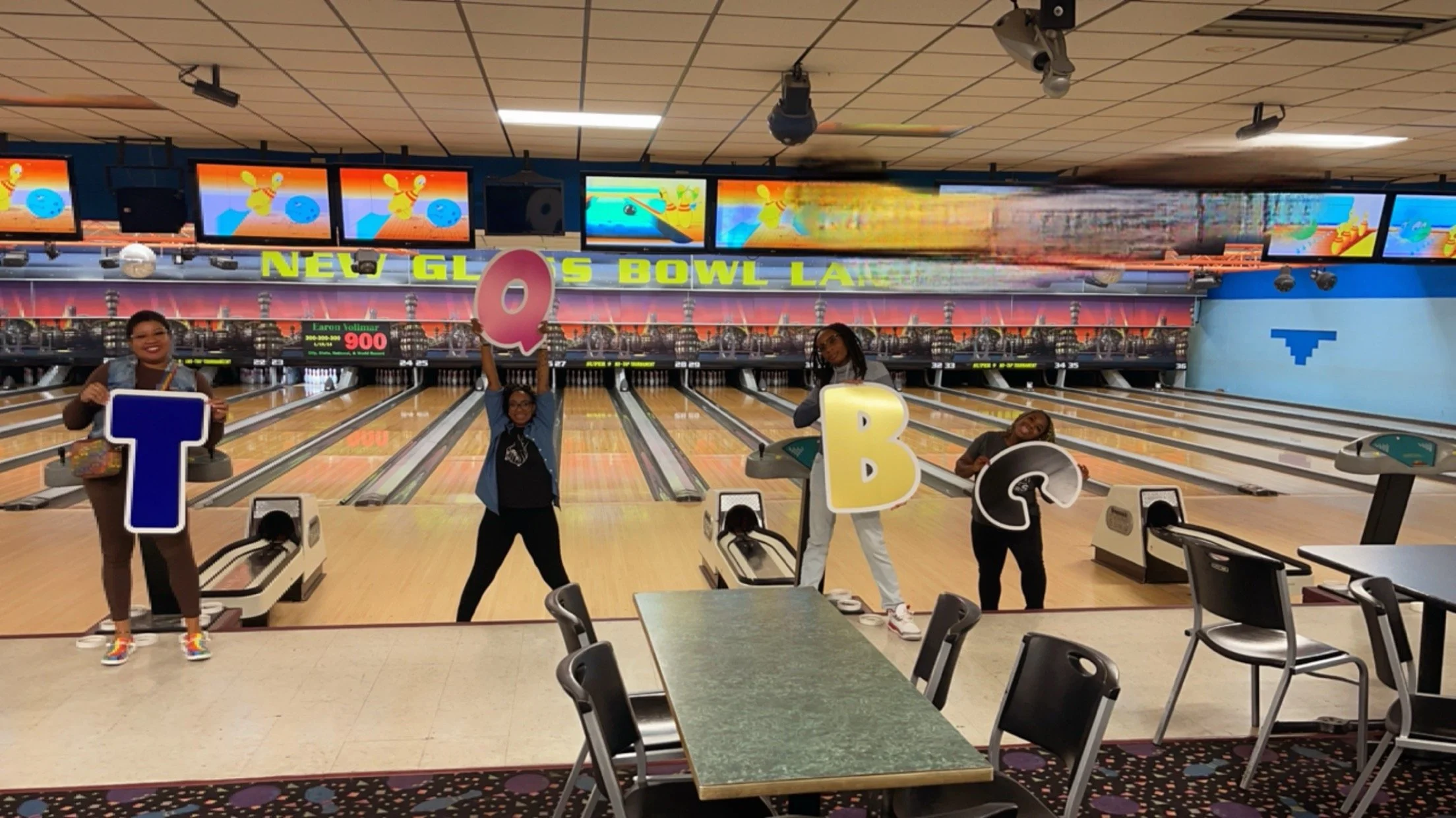 Four women at a bowling alley holding large colorful letter signs spelling TQBB. The bowling lanes are in the background with digital scoreboards and colorful screens above.