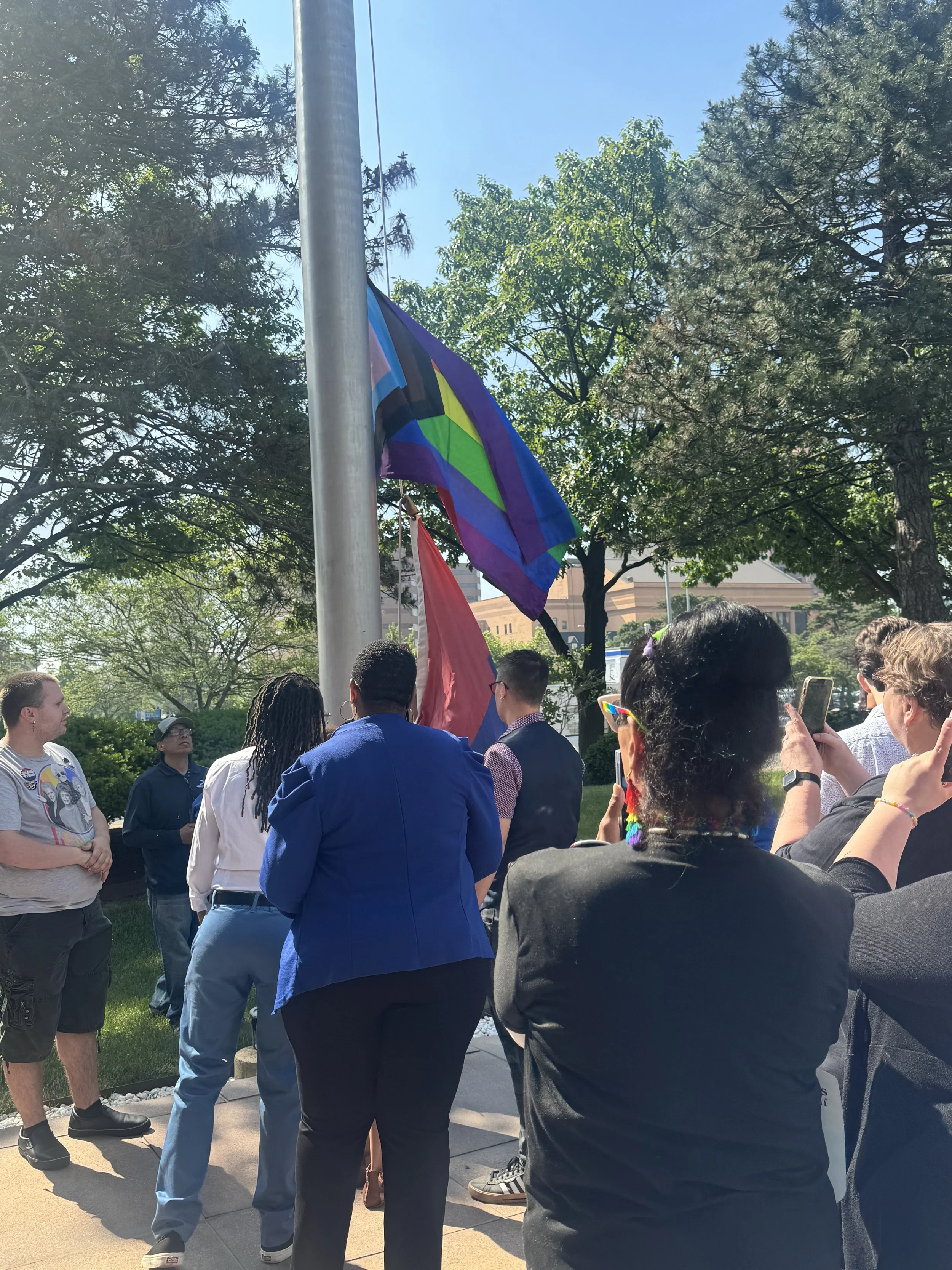 People gathered outdoors near a flagpole with two flags, including a rainbow pride flag, with trees and a building in the background.