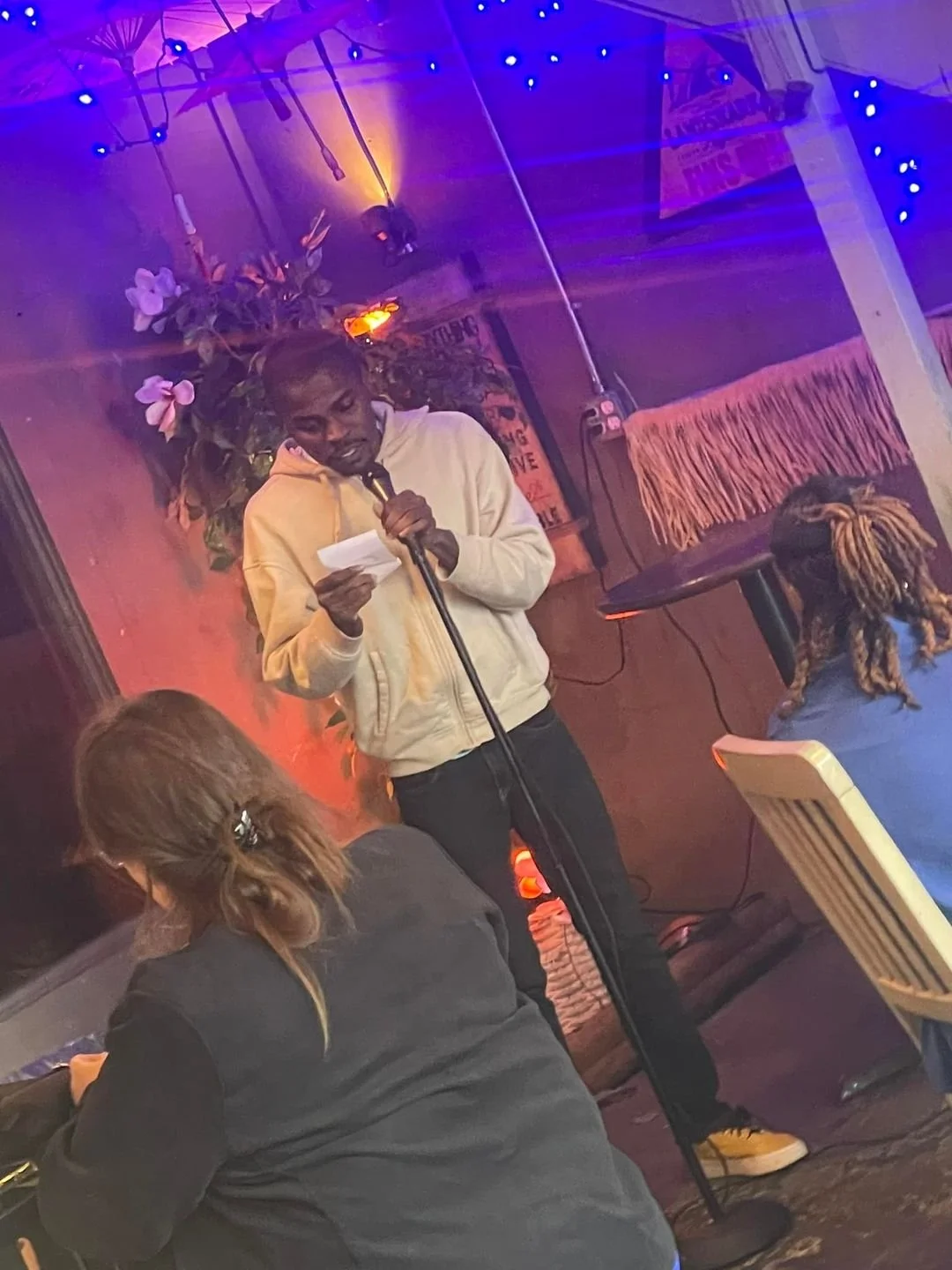 A man with dreadlocks and a yellow hoodie is standing on stage reading from a piece of paper while holding a microphone. Two women are seated, one with glasses and long hair and another with blond dreadlocks, listening. The stage features colorful li