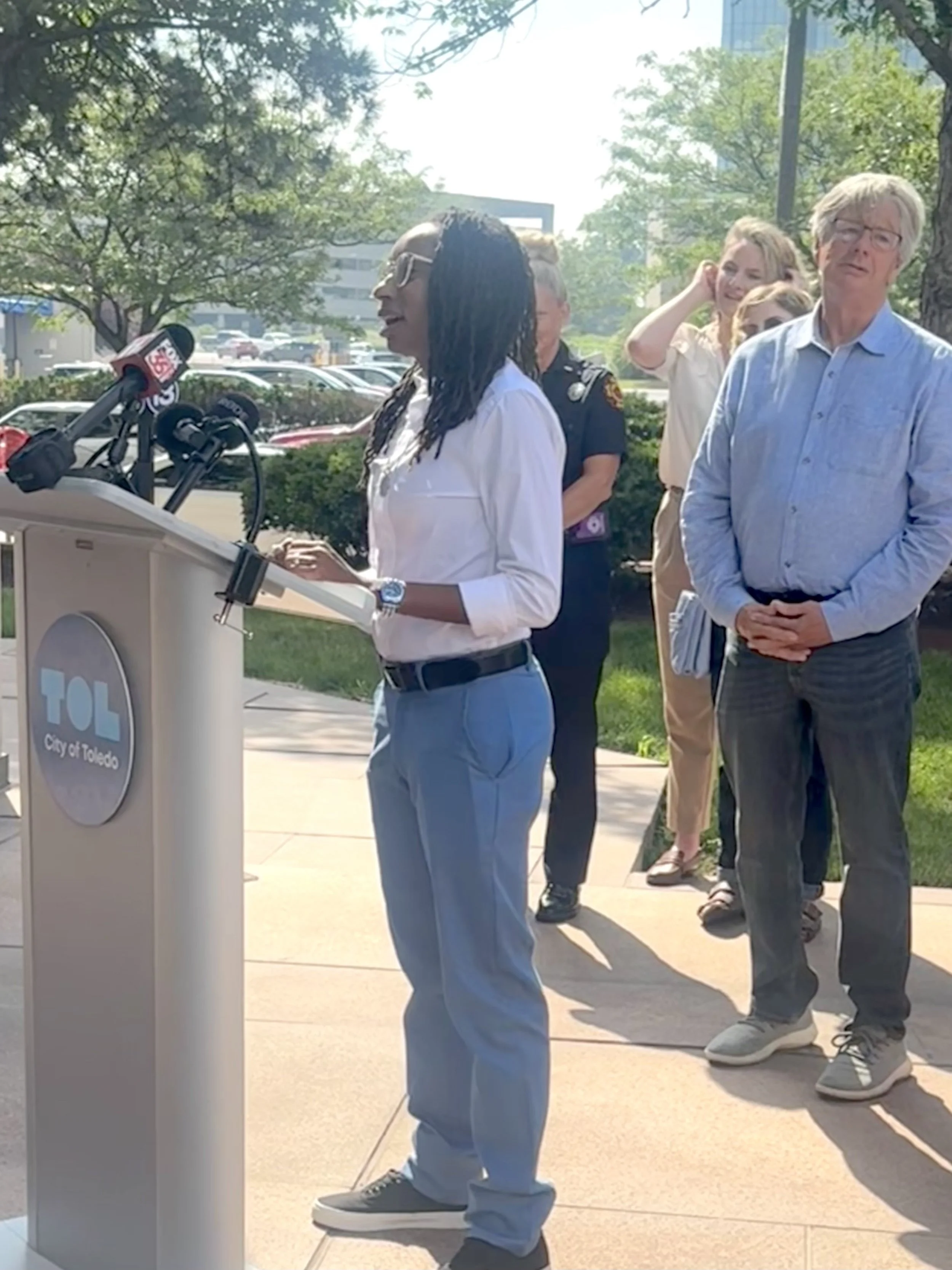 A woman speaking at a podium outdoors with a 'City of Toledo' sign, with several people standing behind her, including a man in a blue shirt and a police officer, on a sunny day with trees and buildings in the background.