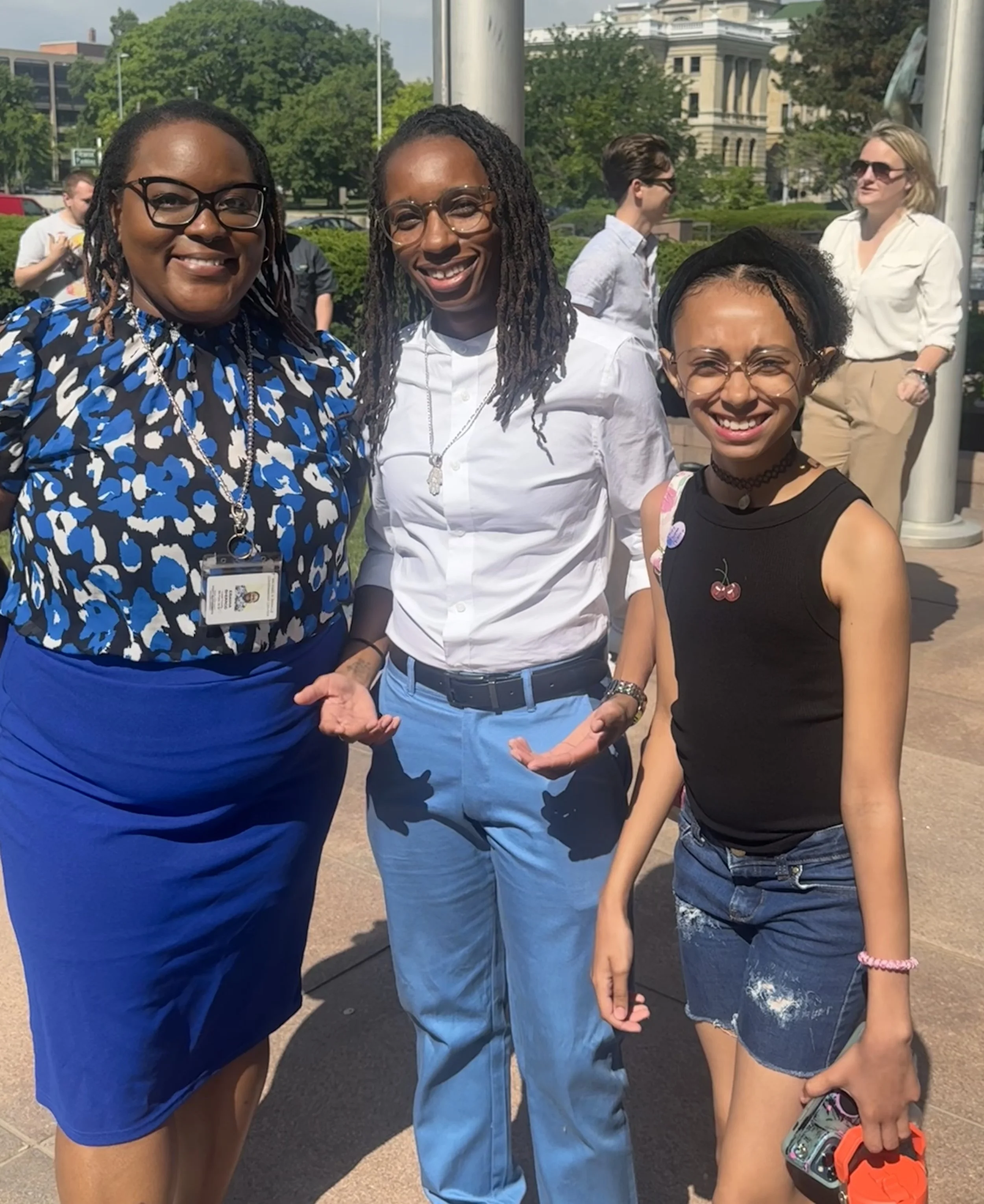 Three women standing outdoors, smiling at the camera, with other people in the background, trees, and buildings on a sunny day.