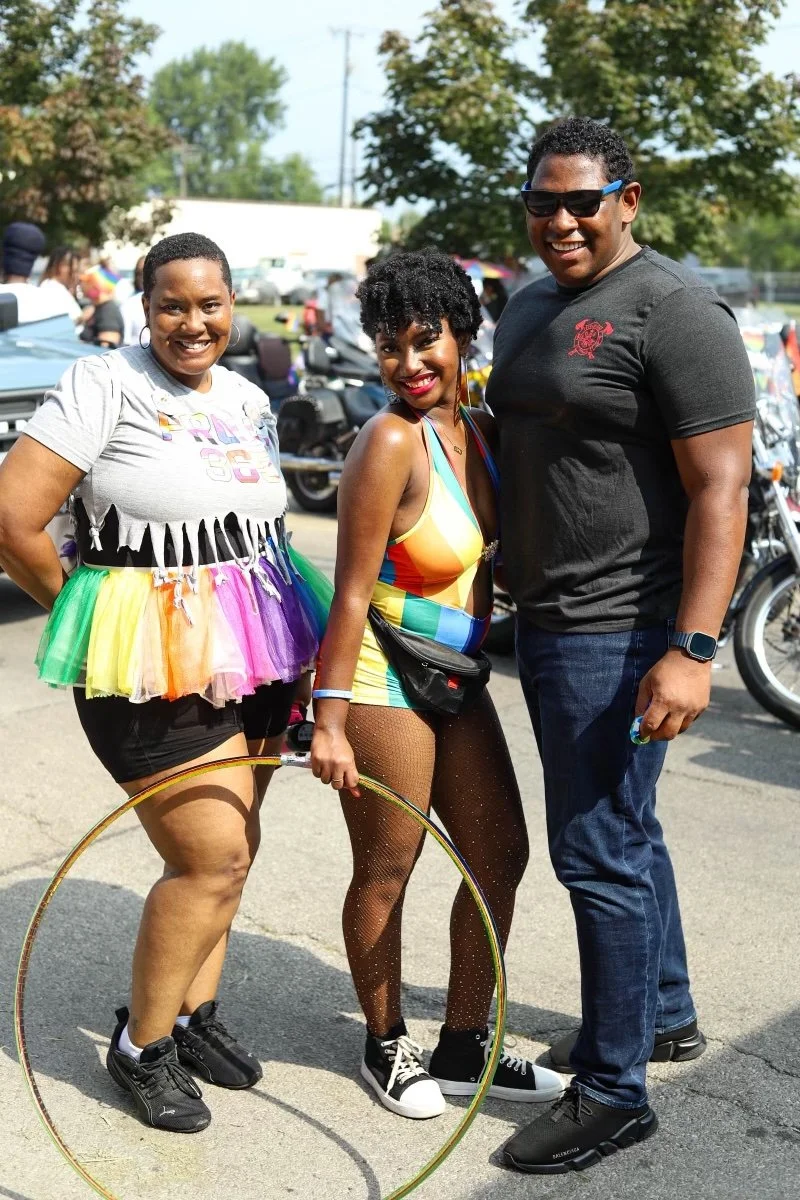 Three smiling people standing outdoors in a parking lot during a sunny day, with motorcycles and trees in the background. One woman is wearing a rainbow tutu and black shorts, another woman is in colorful rainbow attire and fishnet stockings, and a m