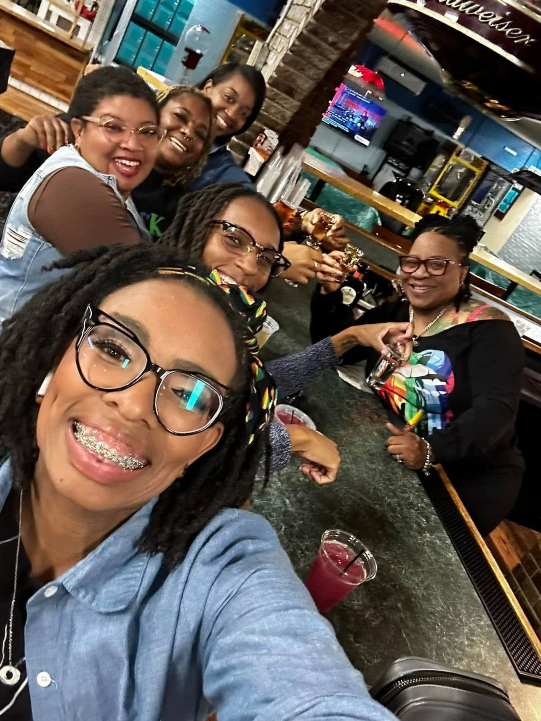 A group of women smiling and sitting at a bar counter, enjoying drinks in a lively bar or restaurant.