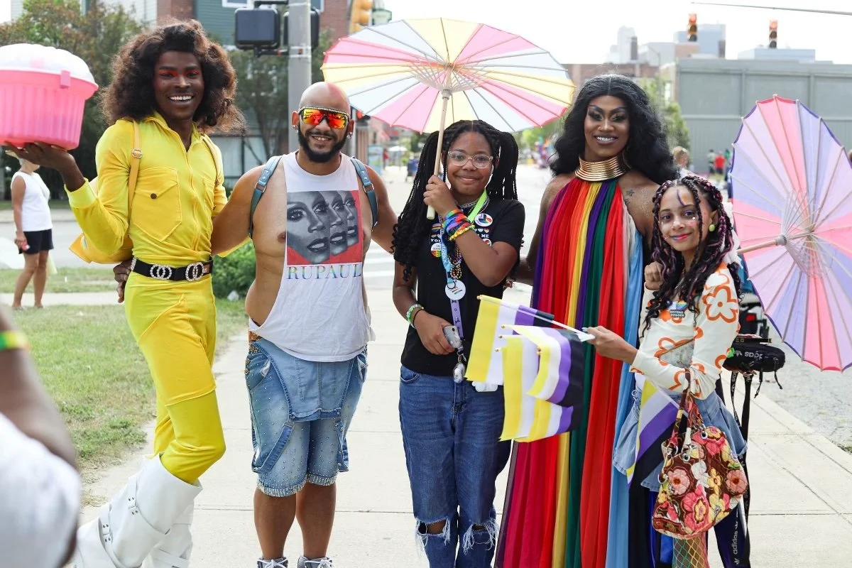 Group of five diverse people celebrating at a Pride parade, holding rainbow and Pride flags, flowers, and umbrellas, dressed in colorful clothing.