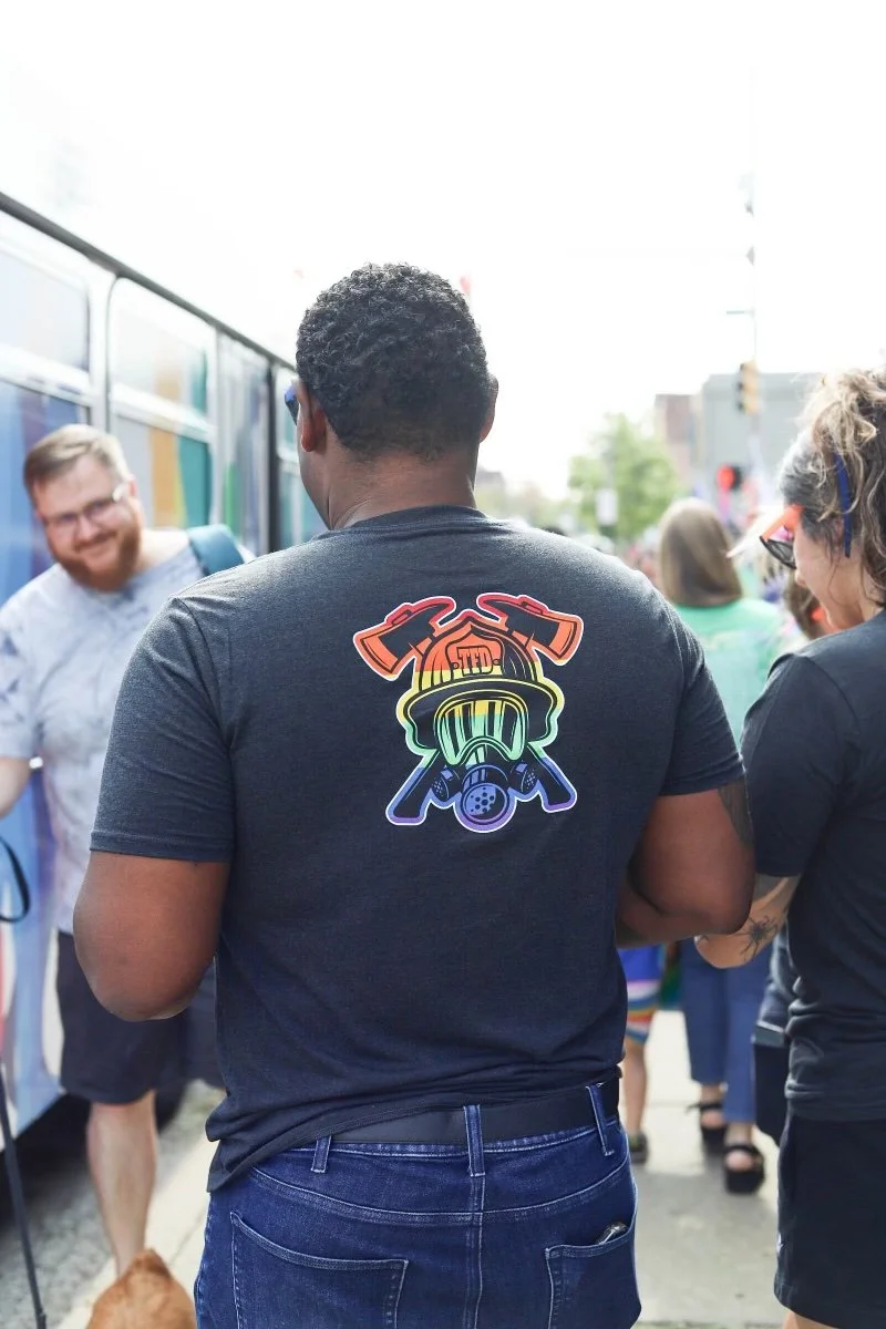 Rear view of a man wearing a black t-shirt with a colorful neon-style design of a gas mask and tools, standing in a crowd outdoors.