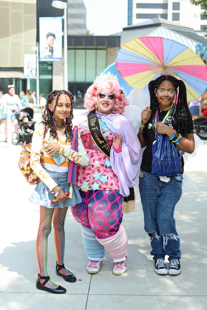 Three people in colorful, whimsical outfits stand together outdoors in an urban area, with buildings and other people in the background. The central person is dressed in a vibrant, playful costume with pink hair, makeup, and a sash, holding an umbrel