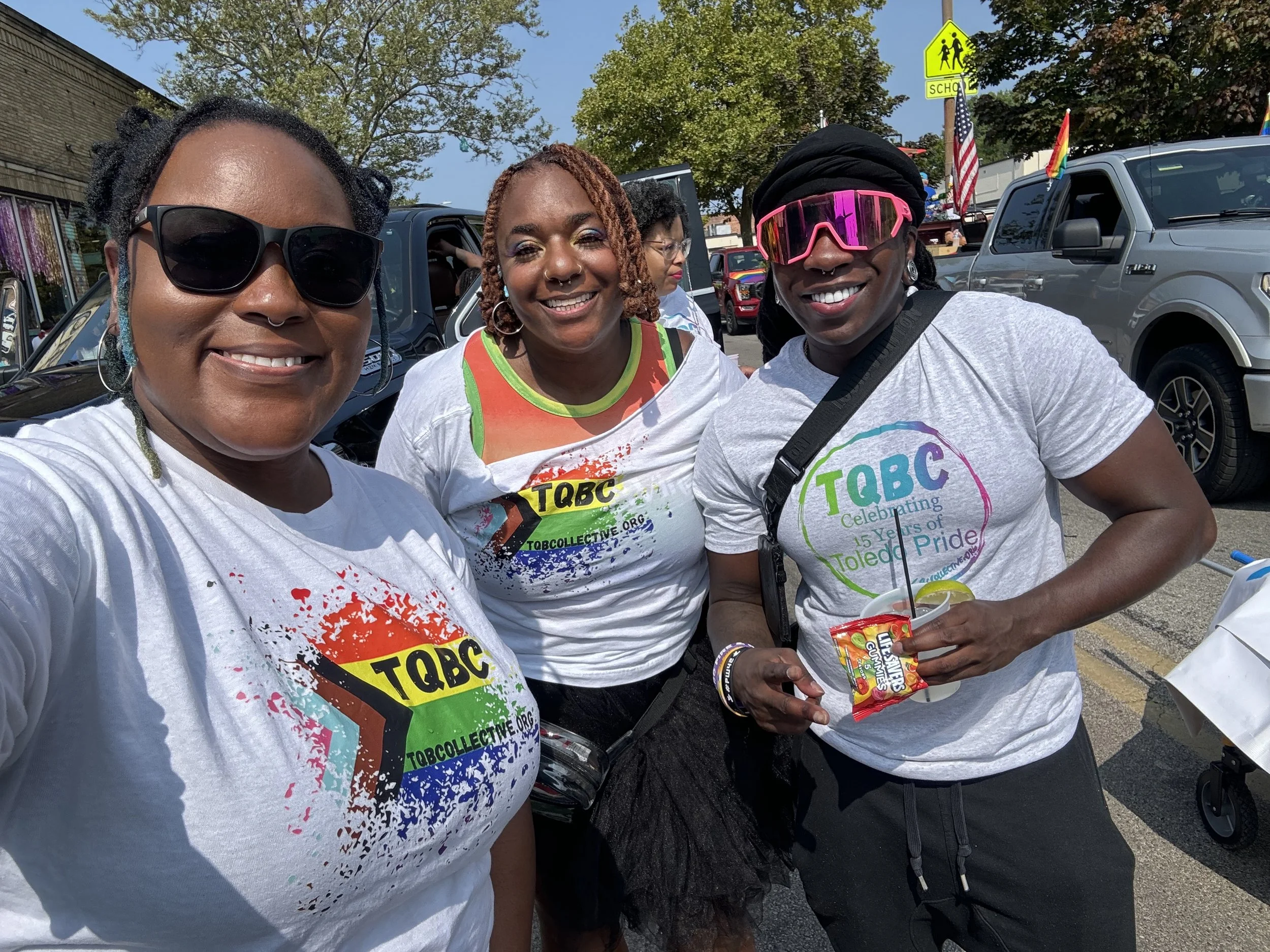 Three women smiling, wearing TQBC T-shirts, at a pride event. One woman has sunglasses, another with pink sunglasses, and third with earrings. They are outdoors with cars in the background and a yellow school zone sign.