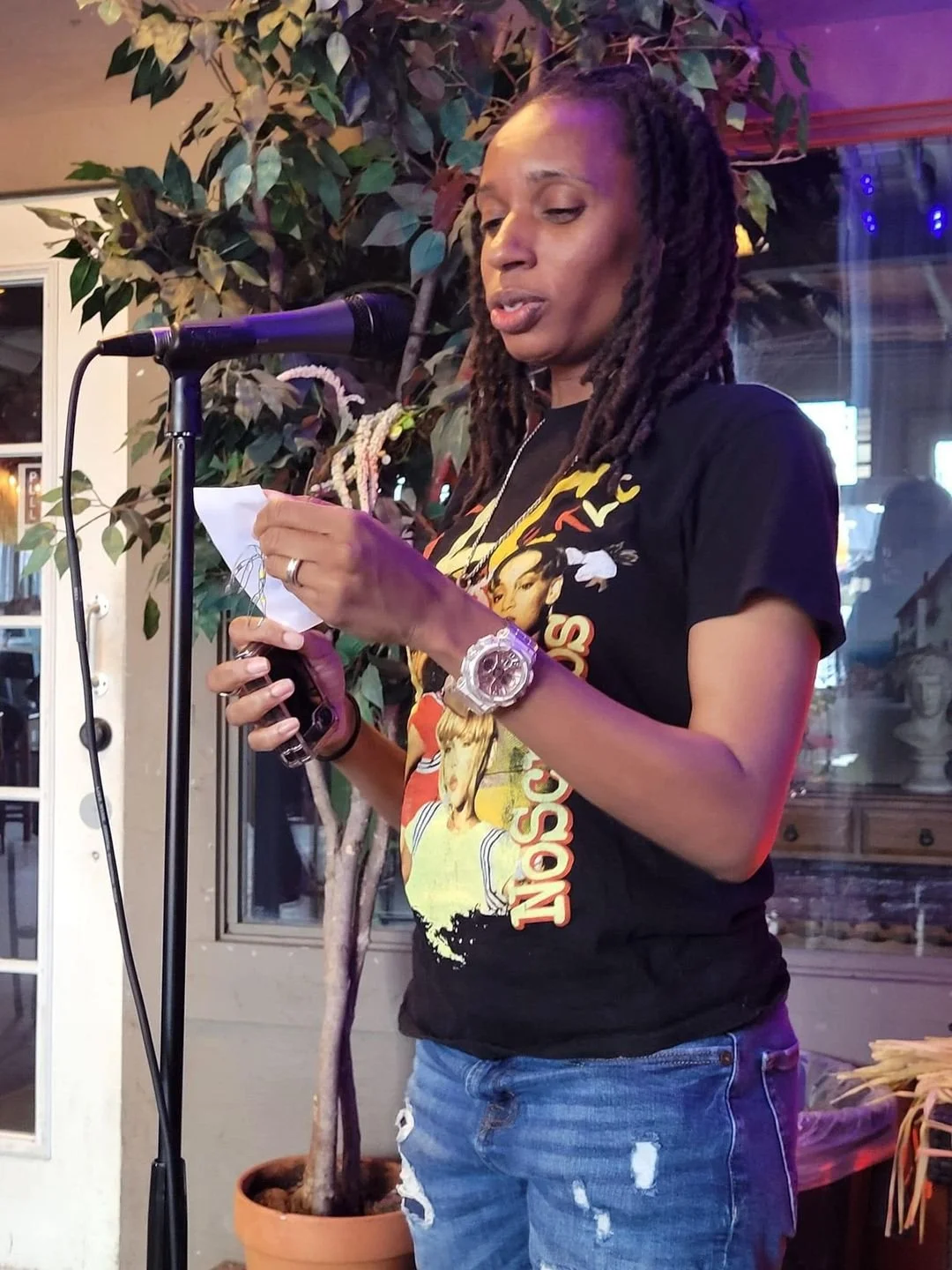 A woman with shoulder-length braided hair, wearing a black T-shirt with a colorful design and distressed jeans, standing in front of a microphone, reading from a piece of paper in a decorated indoor setting with green potted plants.