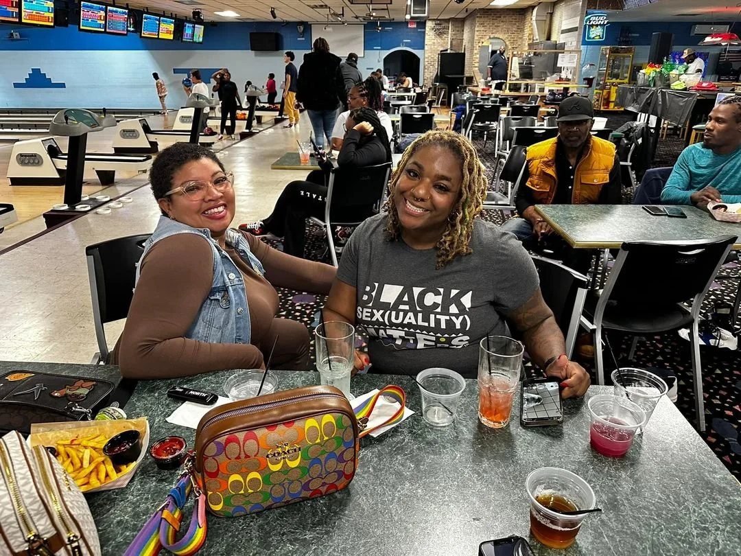 Two women sitting at a bowling alley table, smiling at the camera, with drinks, food, and a colorful purse on the table, and other people and bowling lanes in the background.