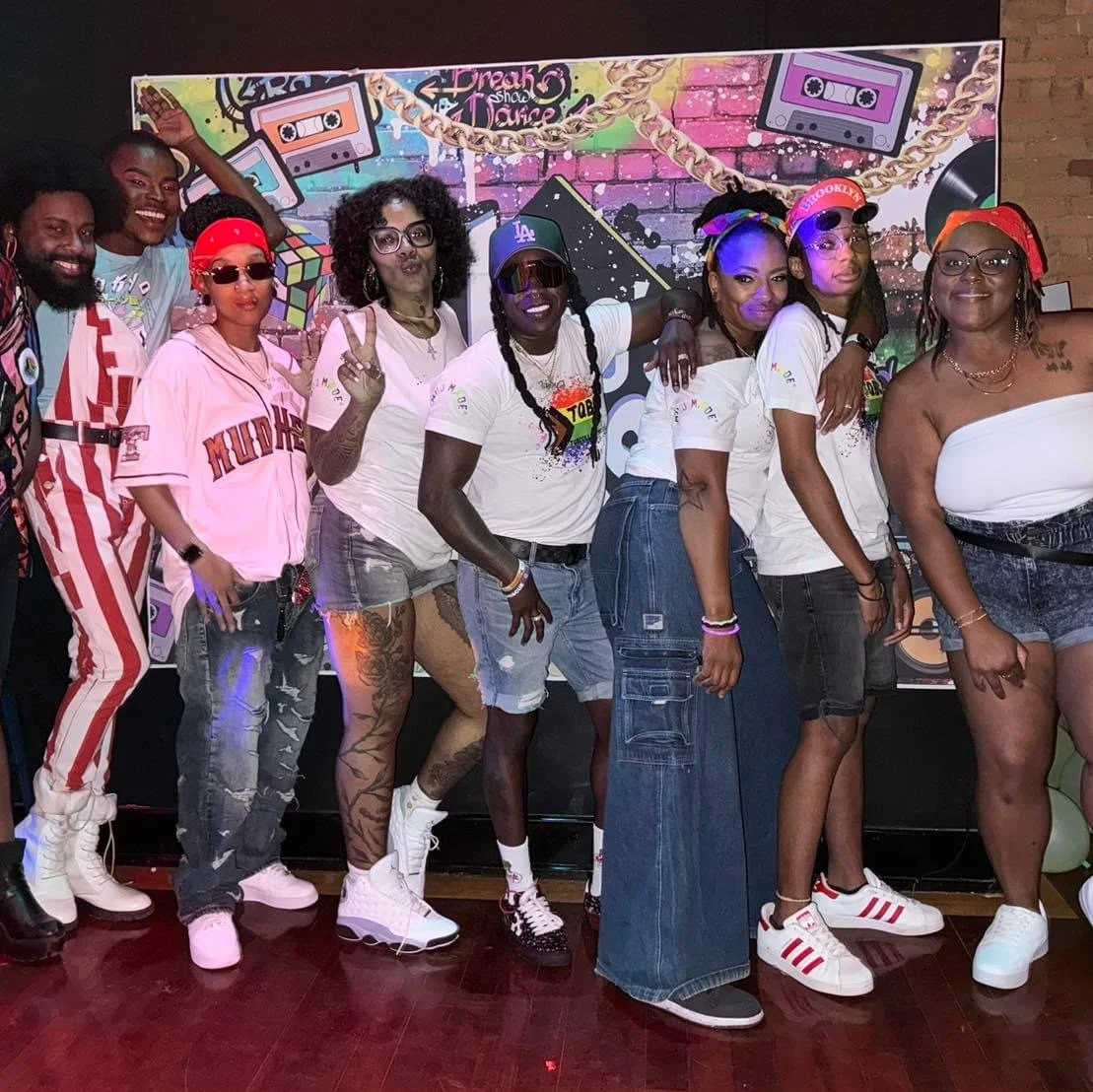 Group of nine people in colorful outfits posing together in front of a graffiti-style backdrop with cassette tapes and musical instruments at a party.