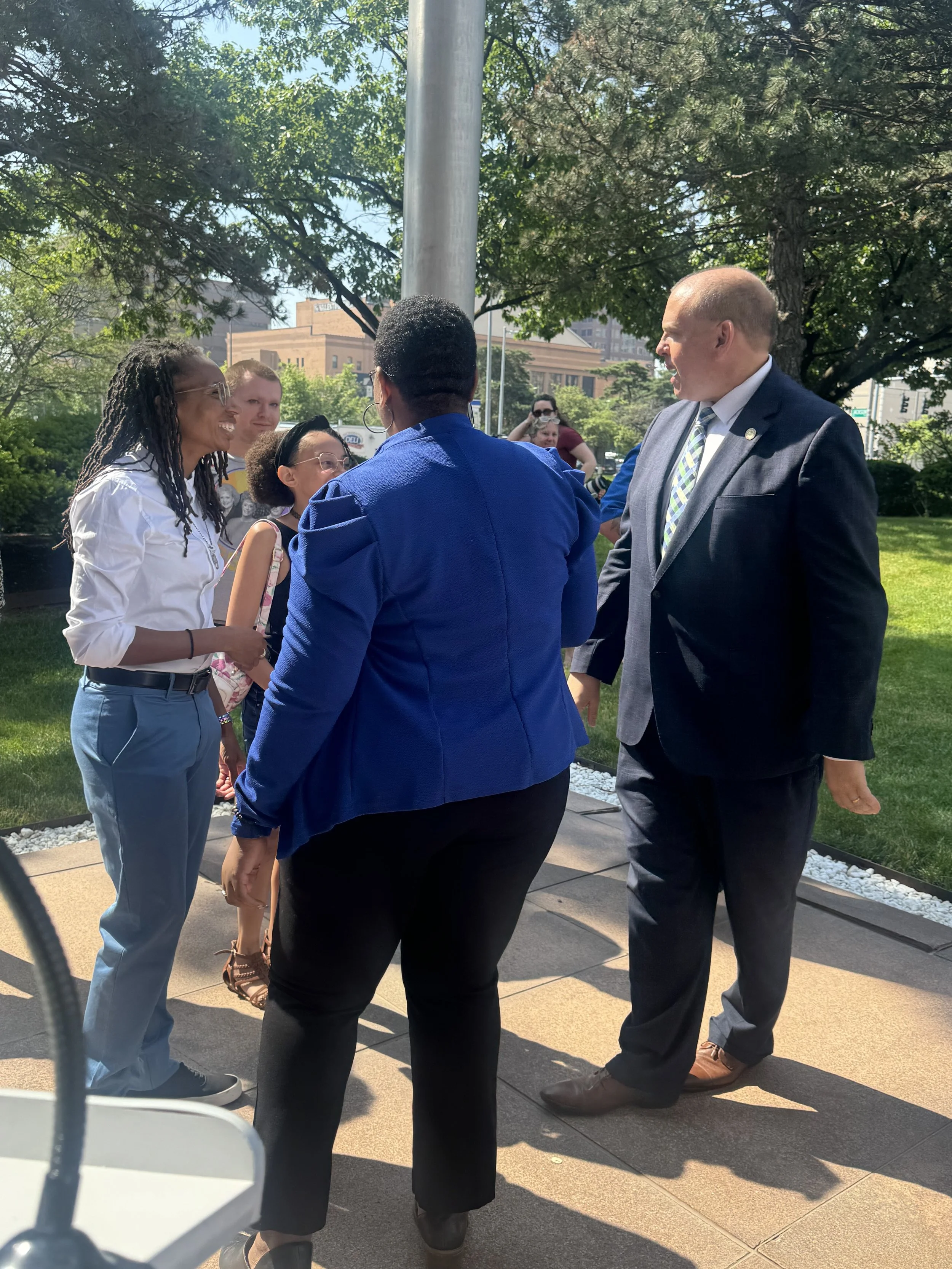 Group of people, including a woman with dreadlocks, a woman in a blue blazer, and a man in a suit, engaging in conversation outdoors on a sunny day, with trees and buildings in the background.